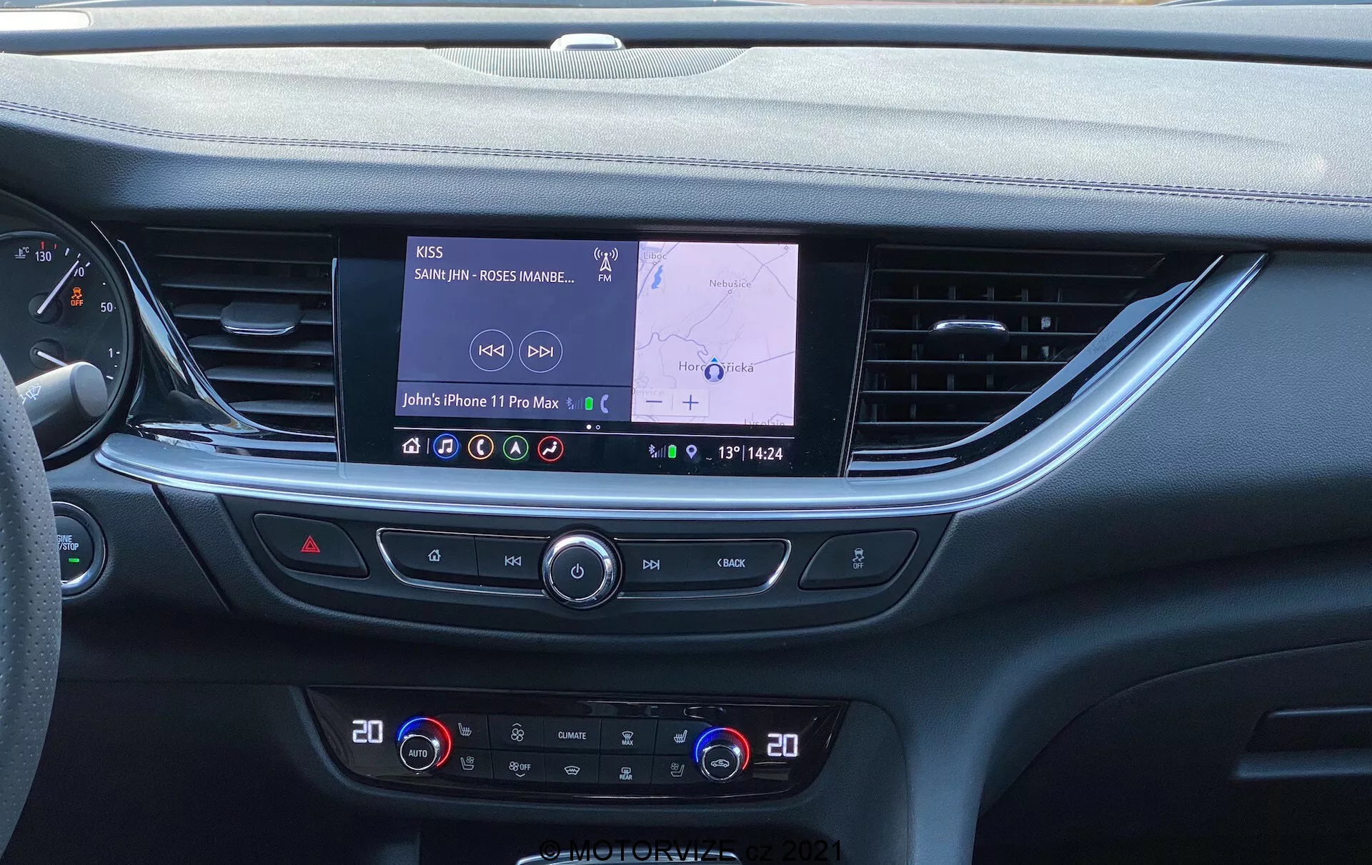 L'intérieur d'une Opel Insignia GSI Grand Sport 2020 moderne, présentant la console centrale et le système d'infodivertissement d'une perspective au niveau des yeux. Une grande écran tactile rectangulaire avec un itinéraire GPS actif et une connexion audio Bluetooth domine la console, entourée de boutons et de cadrans de contrôle de la climatisation. Le design élégant du tableau de bord présente des matériaux au toucher doux, des lignes horizontales et s'étend dans les panneaux de porte. Sous les commandes de climatisation, des boutons supplémentaires pour des fonctions telles que le dégivrage de la lunette arrière, la recirculation de l'air de l'habitacle et le système start/stop sont visibles, avec un espace de rangement ou un chargeur sans fil en bas.