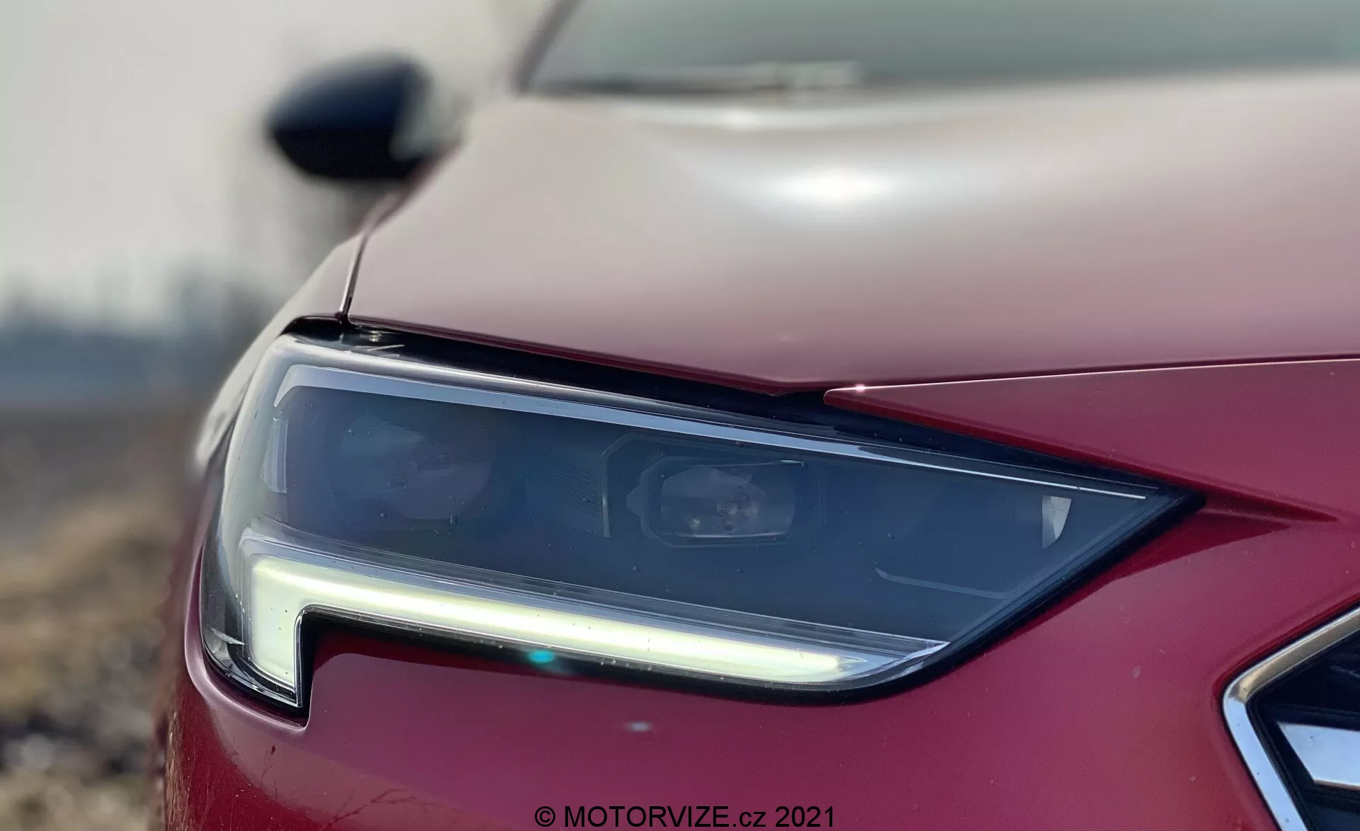 Vue rapprochée de l'ensemble des phares de l'Opel Insignia 2020, se concentrant sur le quart avant gauche du véhicule. Le phare au design moderne présente des lentilles claires avec une technologie LED ou HID, produisant une signature lumineuse angulaire et nette. La photographie met en valeur le design précis du phare et comprend des accents chromés, une vue partielle de la calandre avant et un capot peint en métal, obtenus avec une faible profondeur de champ et un effet bokeh pour une touche artistique.
