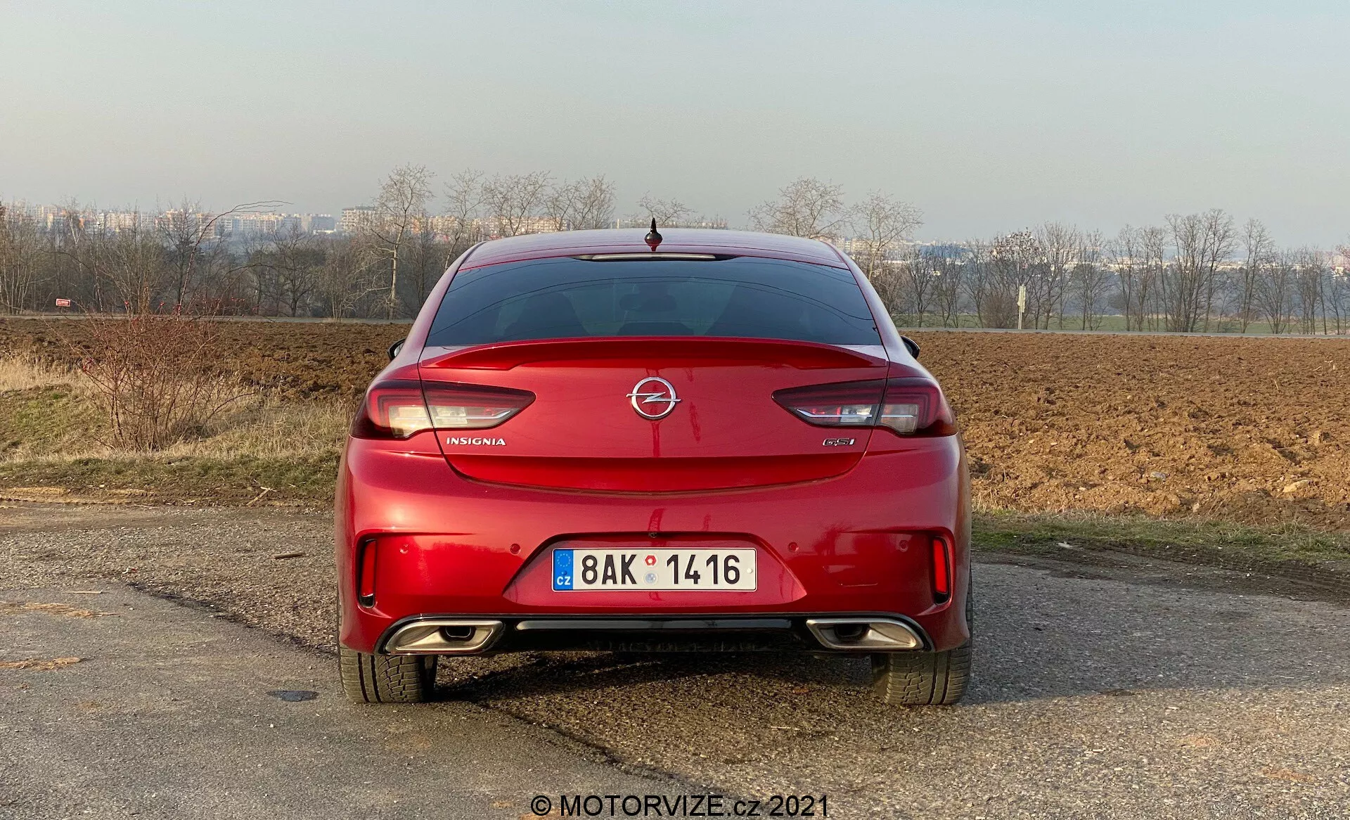 Vue arrière d'une Opel Insignia GSi Grand Sport 2020 rouge garée sur une surface non pavée avec un paysage rural en arrière-plan. La photographie, prise en fin d'après-midi sous une lumière naturelle, montre l'emblème de la marque prominemment sur le couvercle du coffre, avec le nom du modèle en dessous. Le feu stop monté en hauteur est visible sous la lunette arrière. Les feux arrière ont un design courbé autour des côtés du véhicule, caractéristique du style moderne. Le pare-chocs arrière inclut des réflecteurs et possiblement des capteurs de stationnement, avec un système d'échappement double indiquant un design sportif. La plaque d'immatriculation avec un numéro d'enregistrement visible est montée au centre du pare-chocs. Un petit aileron est présent au bord du coffre.