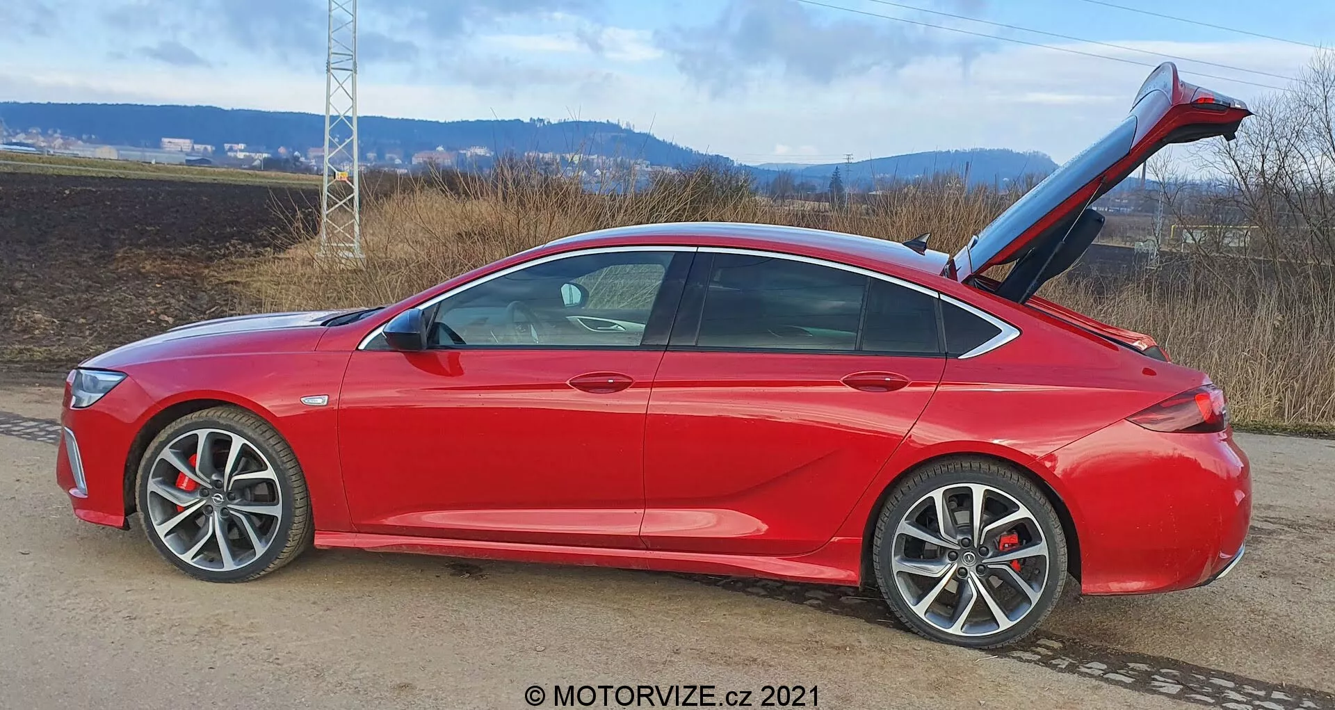 Berline moderne Opel Insignia-GSI-Grand Sport 2020 rouge vif, vue de profil latéral, orientée vers la droite, sur une route avec un paysage naturel; ses caractéristiques comprennent des lignes aérodynamiques, un toit incliné, un coffre ouvert avec hayon automatique, de grandes roues en alliage à plusieurs rayons, des rétroviseurs de couleur carrosserie avec clignotants intégrés, des freins à disque visibles avec étriers, des vitres légèrement teintées, une peinture métallisée, des lignes de caractère définies et une technologie avancée d'ouverture des portes.