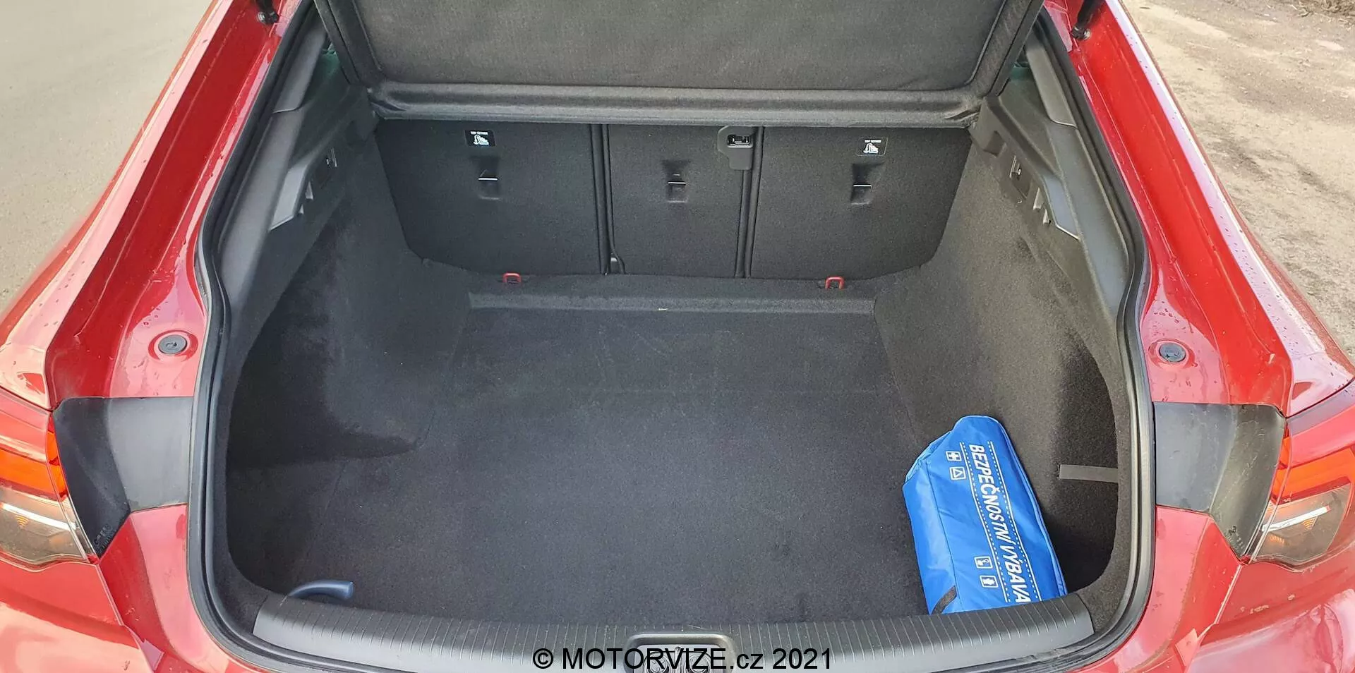 Plan en plongée du coffre ouvert d'une Opel Insignia GSi Grand Sport rouge 2020, vue de l'arrière de la voiture. Le coffre spacieux présente un plancher sombre et capitonné et des dossiers de sièges arrière rabattables 60/40 en position verticale. Des contours et découpes sur les côtés accueillent des éléments structurels, maximisant l'espace utilisable. Un seul objet bleu sur le côté droit sert de point de référence pour la taille.