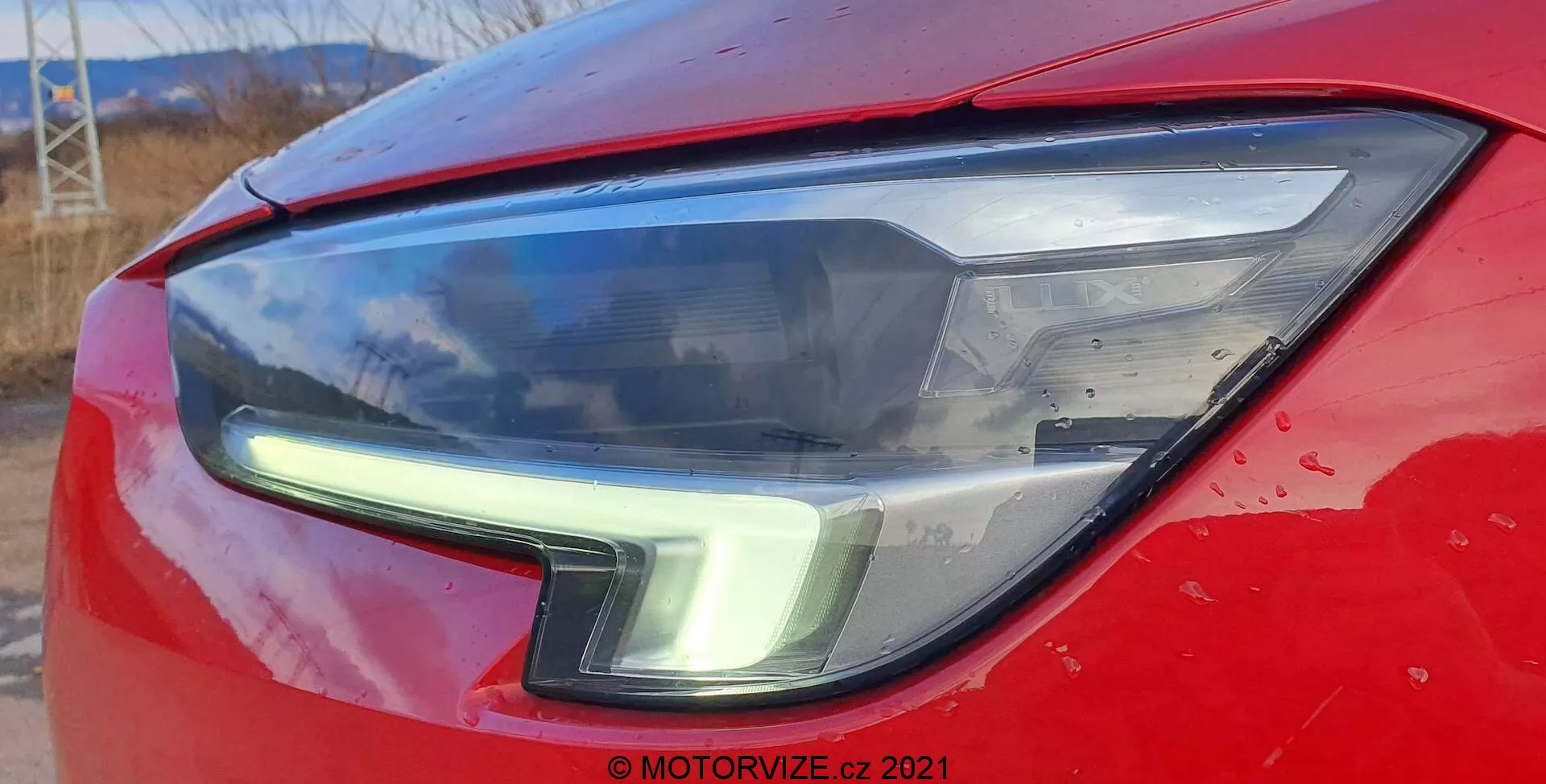 Gros plan sur l'unité de phare avant gauche de l'Opel Insignia 2020 GSi Grand Sport, présentant un design moderne et élégant avec des feux diurnes à LED se courbant vers le haut le long du bord extérieur. Le capot, l'aile et le boîtier de phare transparent de couleur rouge vif soulignent le design aérodynamique et la technologie d'éclairage contemporaine de la voiture. De petites taches d'eau et de la saleté indiquent que la voiture est utilisée régulièrement.