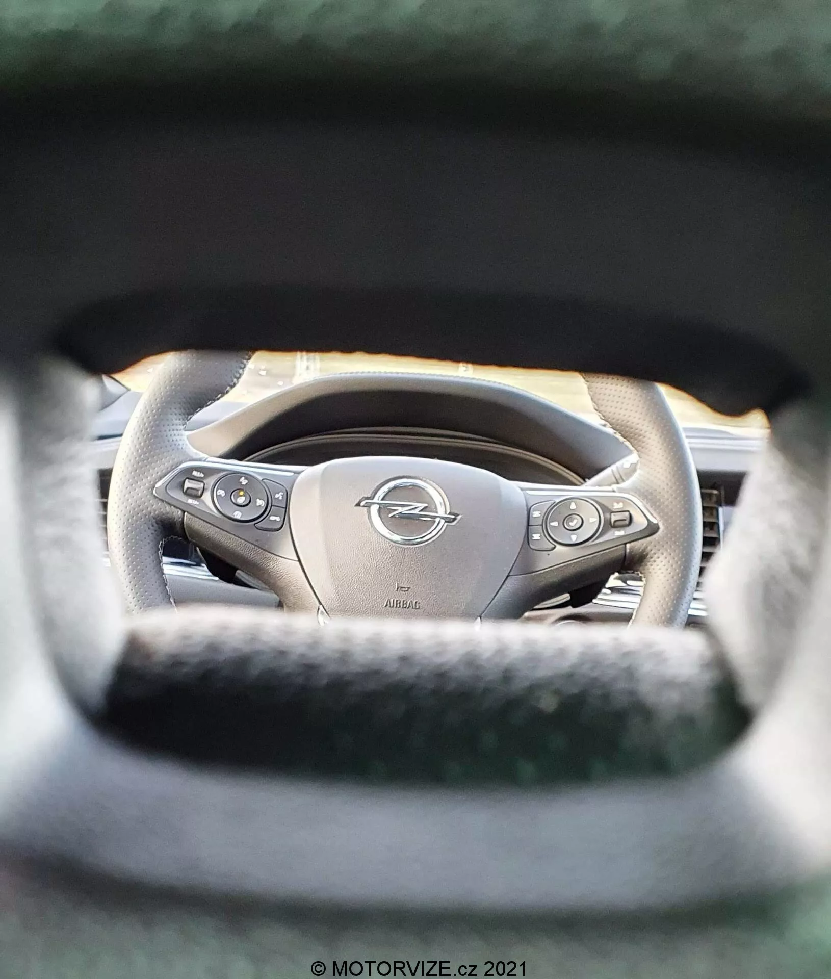 Intérieur de l'Opel Insignia 2020 (Insignia GSi Grand Sport) avec une vue détaillée du volant depuis la perspective du siège conducteur. Le point focal est le volant centré sur l'emblème Opel, d'un design ergonomique et revêtu de cuir avec des boutons multifonctionnels. La faible profondeur de champ met en évidence le volant tout en floutant la zone environnante, y compris les instruments du tableau de bord. Les caractéristiques remarquables incluent des accents métalliques, une possible fonction de chauffage et une gravure 'AIRBAG', soulignant les éléments de luxe et de sécurité.