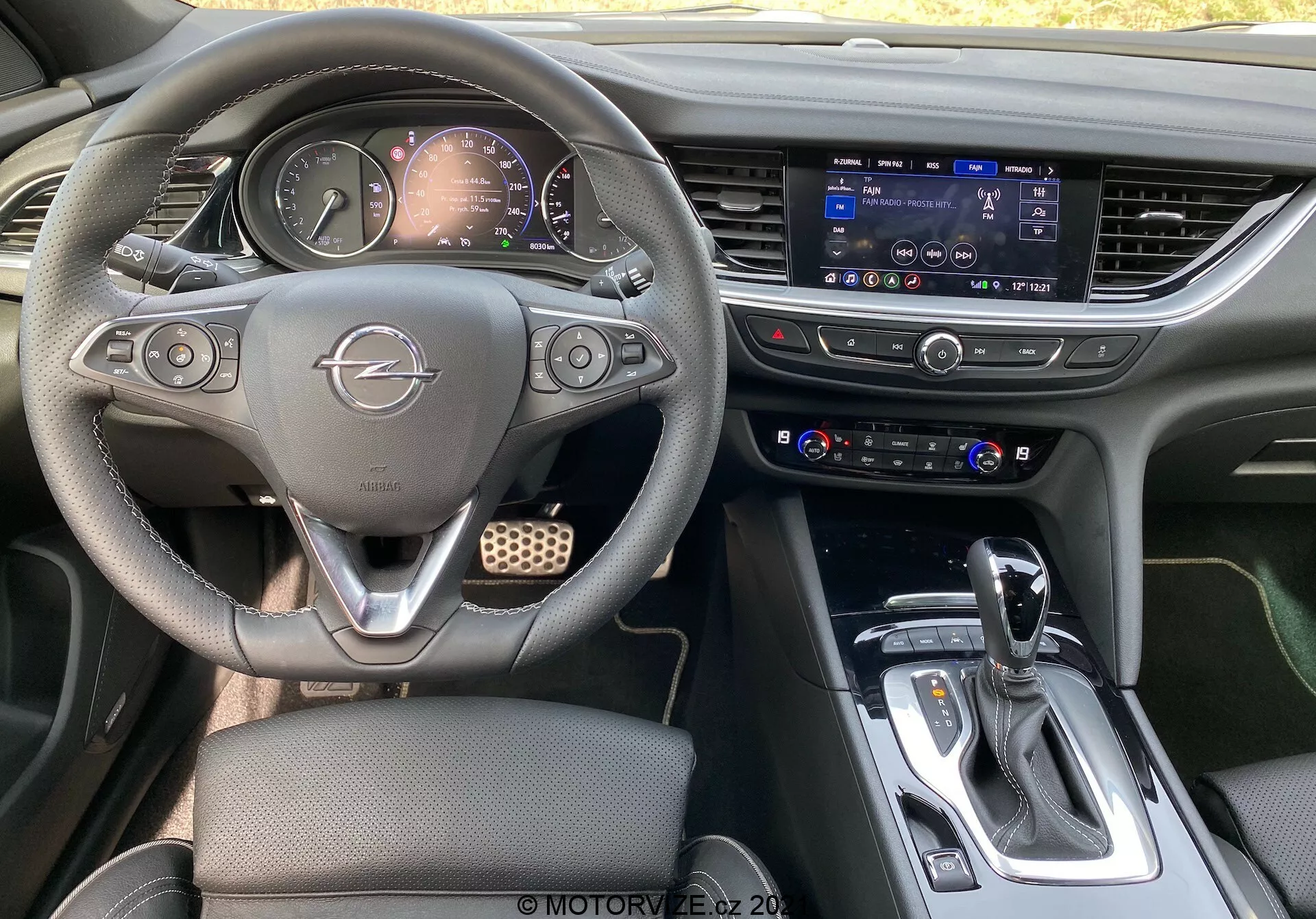 Vue intérieure d'une Opel Insignia GSi Grand Sport 2020 depuis les sièges arrière, montrant le volant multifonctions avec logo Opel, le tableau de bord partiellement visible avec cadrans analogiques et affichage numérique, l'écran tactile proéminent du système d'infodivertissement, le panneau de contrôle du climat, le levier de vitesses automatique avec soufflet en cuir et accents argentés, les porte-gobelets, la console centrale avec espace de rangement et un design intérieur sombre et contemporain avec des accents métalliques, indiquant un véhicule moderne et possiblement haut de gamme.