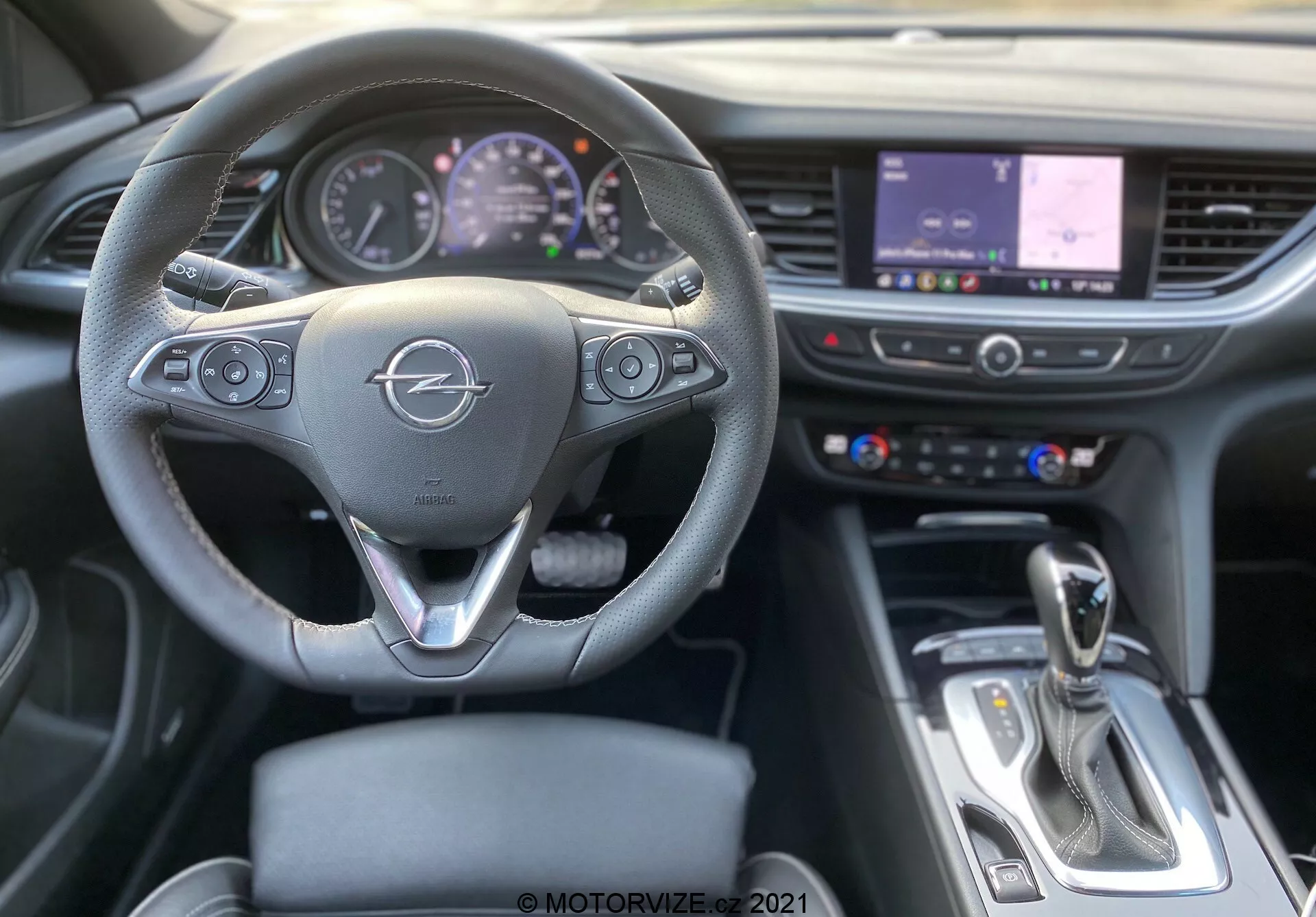 Vue intérieure d'une Opel Insignia GSI Grand Sport 2020 depuis la perspective du siège conducteur, se concentrant sur le volant à trois branches avec boutons de commande multimédia et de véhicule, tableau de bord mixte analogique et numérique, écran tactile d'infodivertissement central affichant une carte, levier de vitesse, accents chromés et un mélange moderne de matériaux comme le cuir et le métal.