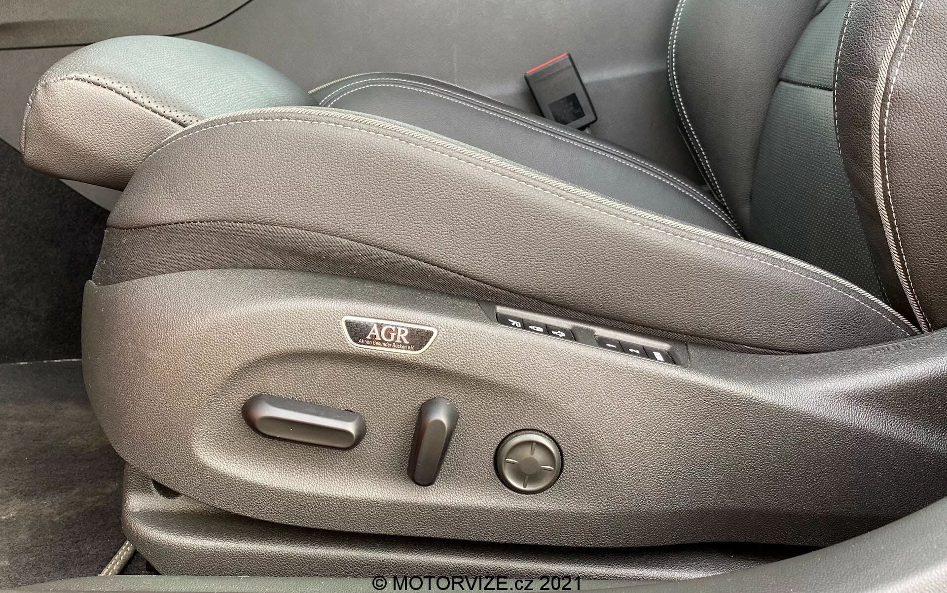 Gros plan sur le côté d'un siège avant dans une Opel Insignia GSi Grand Sport 2020, se concentrant sur les commandes de réglage du siège. L'image capture le revêtement de couleur gris foncé ou noir, les boutons latéraux pour la position avant et arrière, les boutons verticaux pour incliner le dossier, un bouton rond pour le support lombaire, un logo 'AGR' et un levier probablement pour le réglage en hauteur du siège. © MOTORVIZE.cz 2021 filigrane.