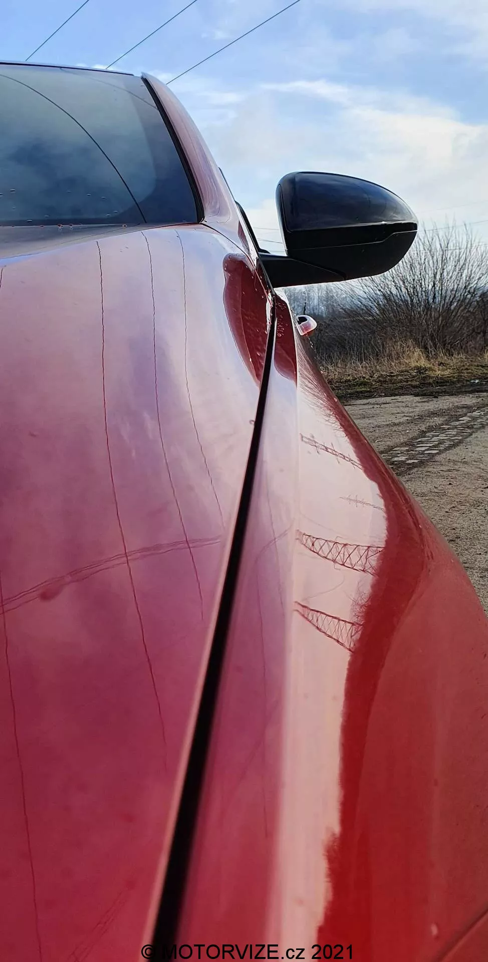 Gros plan vertical de l'Opel Insignia 2020 rouge brillant (Insignia-GSI-Grand-Sport) se concentrant sur le profil latéral près de la porte du conducteur. L'image montre le rétroviseur noir avec clignotant intégré, le bord de la porte avec couture et rayures visibles, et des reflets d'un environnement semi-rural avec un ciel dégagé.
