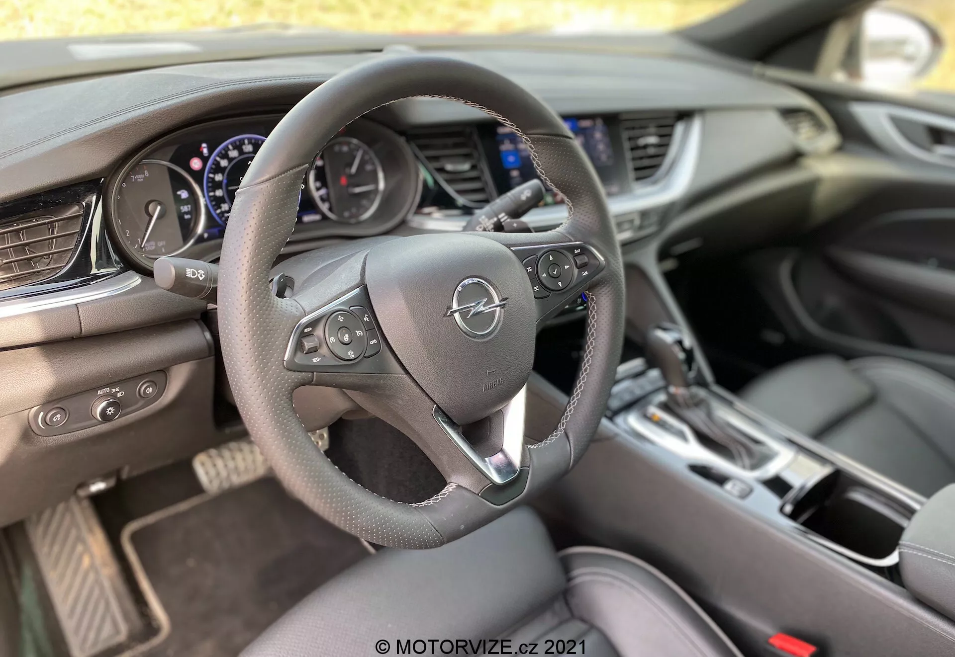 Vue intérieure d'une Opel Insignia GSI Grand Sport 2020 depuis la perspective du conducteur, mettant en évidence un volant en cuir à trois branches avec commandes multifonctions, un combiné d’instruments avec cadrans analogiques et écran numérique central, un grand écran d'infodivertissement sur la console centrale, des aérateurs horizontaux, une interface de contrôle de la climatisation avec boutons physiques, un levier de sélection de vitesses pour une transmission automatique, des porte-gobelets et un sélecteur de mode de conduite sur le tunnel de transmission, le tout éclairé par la lumière naturelle.