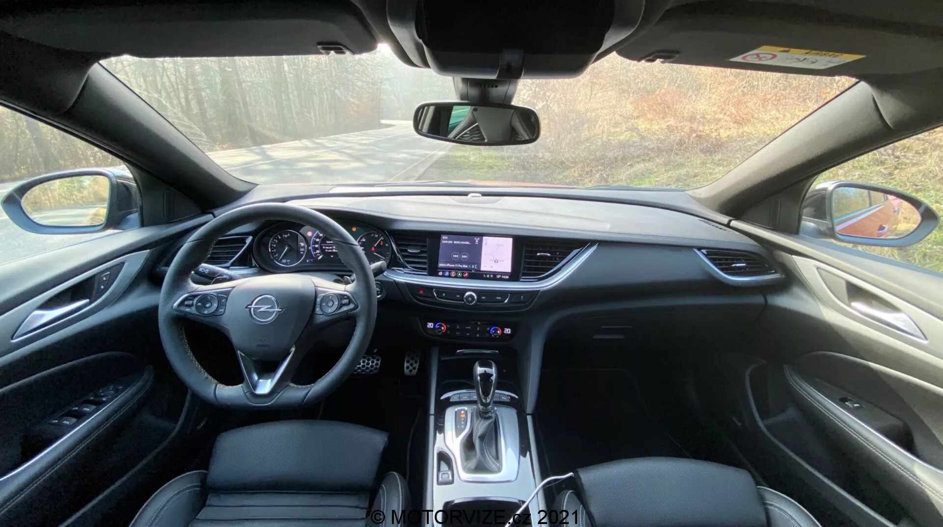 Vue intérieure de l'Opel Insignia GSi Grand Sport 2020 depuis la perspective du conducteur, montrant le tableau de bord avec un volant central équipé de commandes intégrées, un tableau de bord numérique ou traditionnel partiellement visible, une console centrale proéminente avec un système d'infodivertissement à écran tactile, des boutons physiques et des cadrans pour le contrôle climatique sous l'écran tactile, et un sélecteur de transmission automatique. Le design élégant du tableau de bord intègre des plastiques doux au toucher, des pièces de garniture accentuées et une sellerie de haute qualité, le tout baigné par la lumière naturelle du jour.