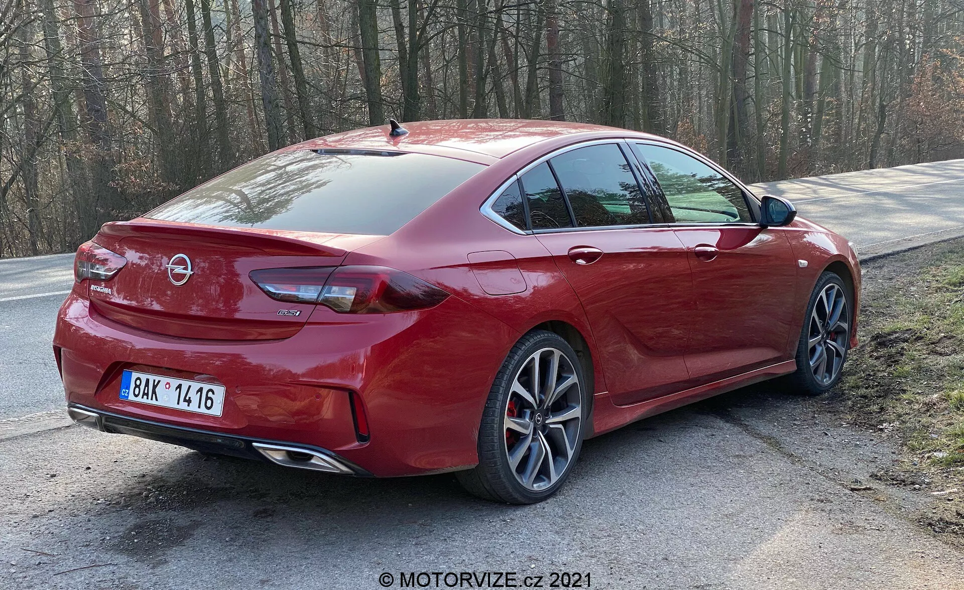 L'image montre une berline rouge Opel Insignia 2020 (Insignia GSi Grand Sport) garée sur le bord de la route au milieu des arbres, suggérant un environnement rural ou ressemblant à un parc. La vue de trois quarts arrière capture le design élégant et sportif de la voiture, incluant le pare-brise arrière incliné, les groupes de feux arrière modernes à LED, les roues en alliage multi-branches, le système d'échappement double, les capteurs de stationnement arrière et l'antenne en forme d'aileron de requin. La garde au sol est standard pour une berline, équilibrant l'utilisation quotidienne avec une configuration orientée vers le sport.