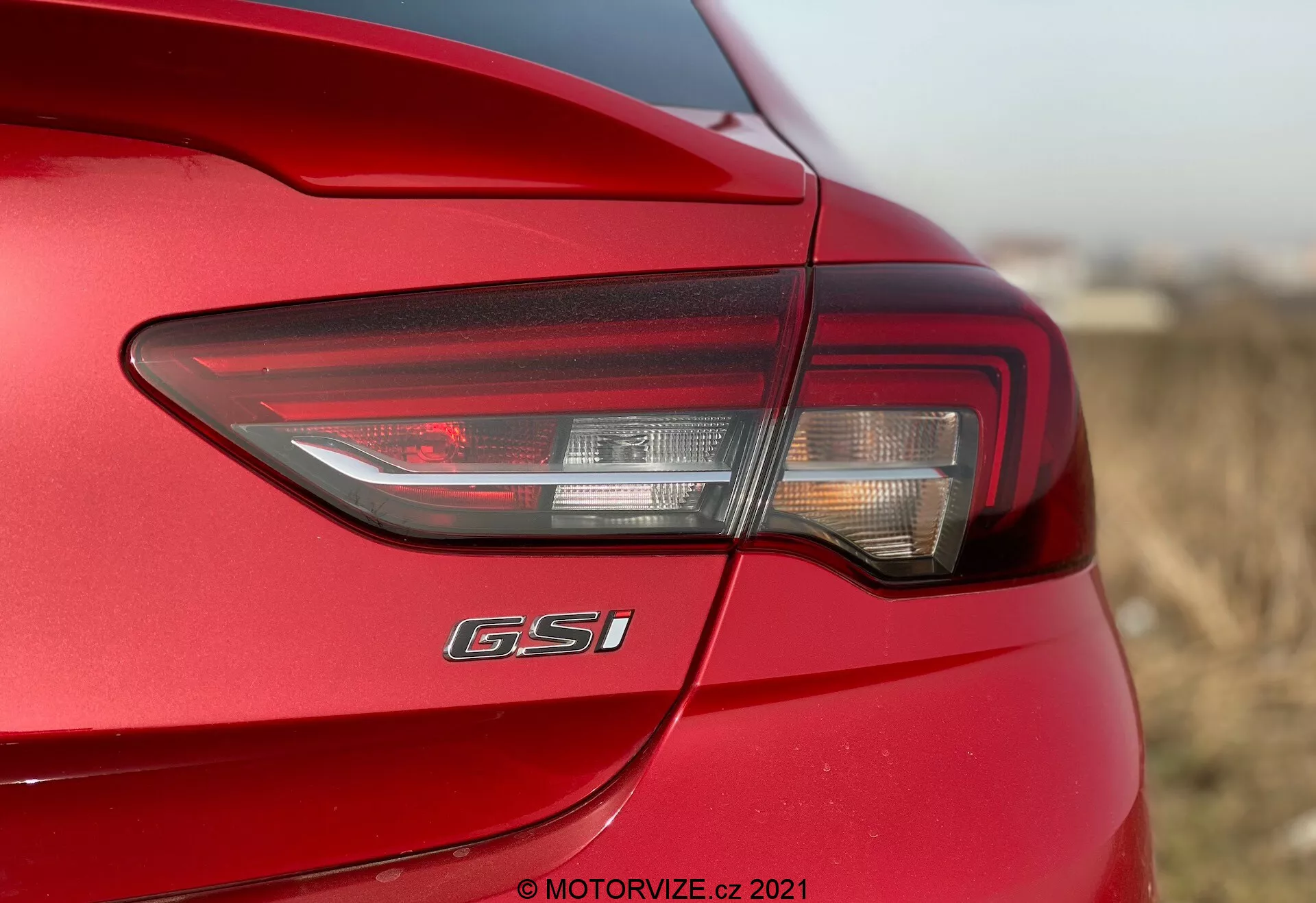 Vue rapprochée du côté arrière gauche de l'Opel Insignia GSi Grand Sport 2020, mettant en avant le design moderne de son feu arrière à LED combinant feux de freinage, clignotants et possiblement feux de recul. Le feu arrière enveloppe l'angle du véhicule, améliorant la visibilité. Sous le feu arrière, l'insigne 'GSi' est clairement affiché, indiquant le niveau de finition du modèle. La voiture arbore une finition de peinture rouge brillante avec des lignes de carrosserie aérodynamiques. La photographie met en avant le feu arrière avec une faible profondeur de champ, soulignant les détails du design tout en floutant les zones environnantes.