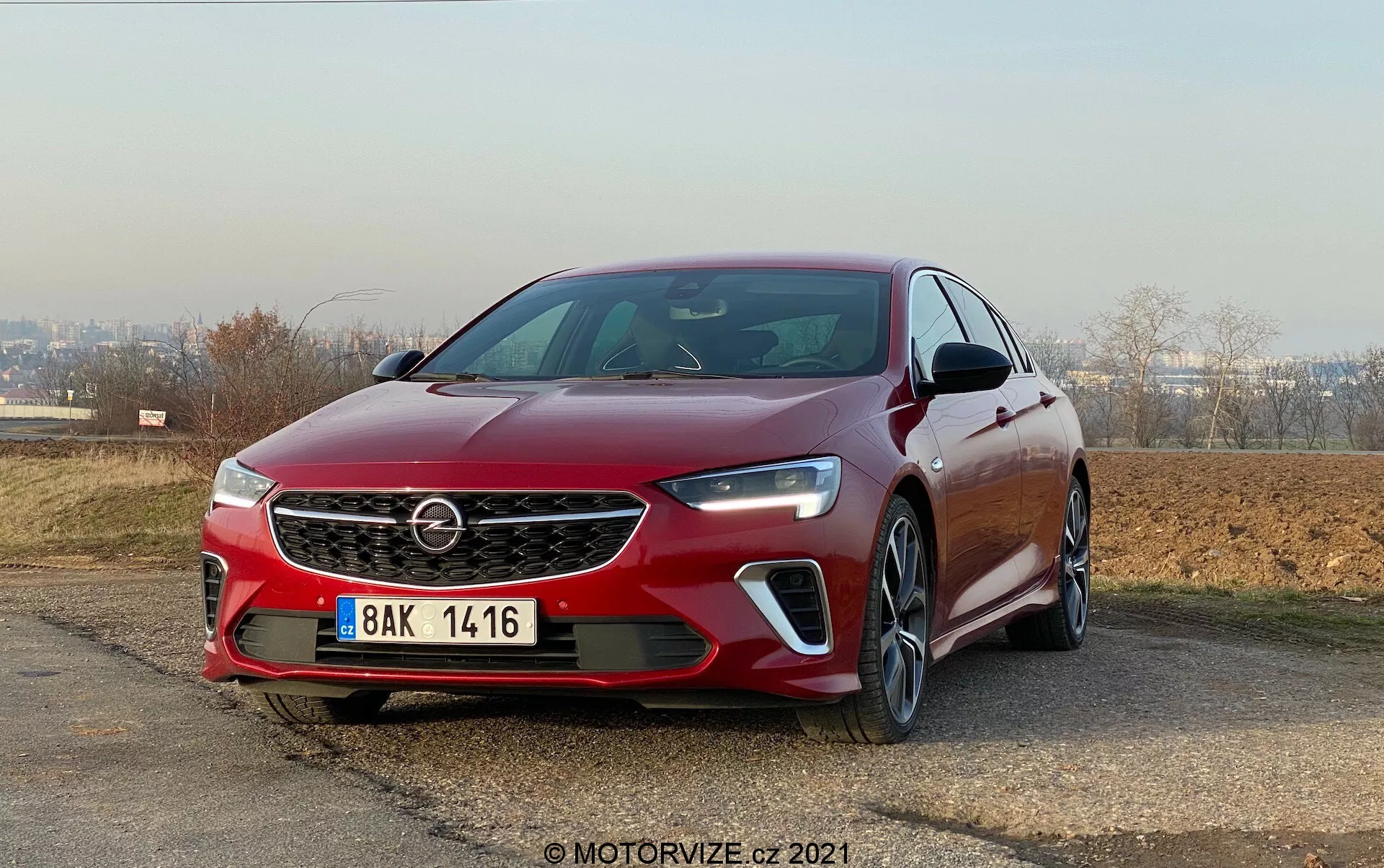 Vue du quart avant gauche d'une Opel Insignia GSi Grand Sport 2020 sous un angle bas, mettant en avant sa calandre proéminente avec logo, ses phares LED angulaires élégants et ses logements de phares antibrouillard avec feux de jour. L'image met en évidence les lignes nettes du capot, le capteur avant sous la plaque d'immatriculation, les lignes sculptées de la carrosserie, les clignotants intégrés dans les rétroviseurs latéraux et les roues en alliage multi-branches. La voiture est positionnée en extérieur avec un ciel dégagé en arrière-plan, garée sur une légère inclinaison.