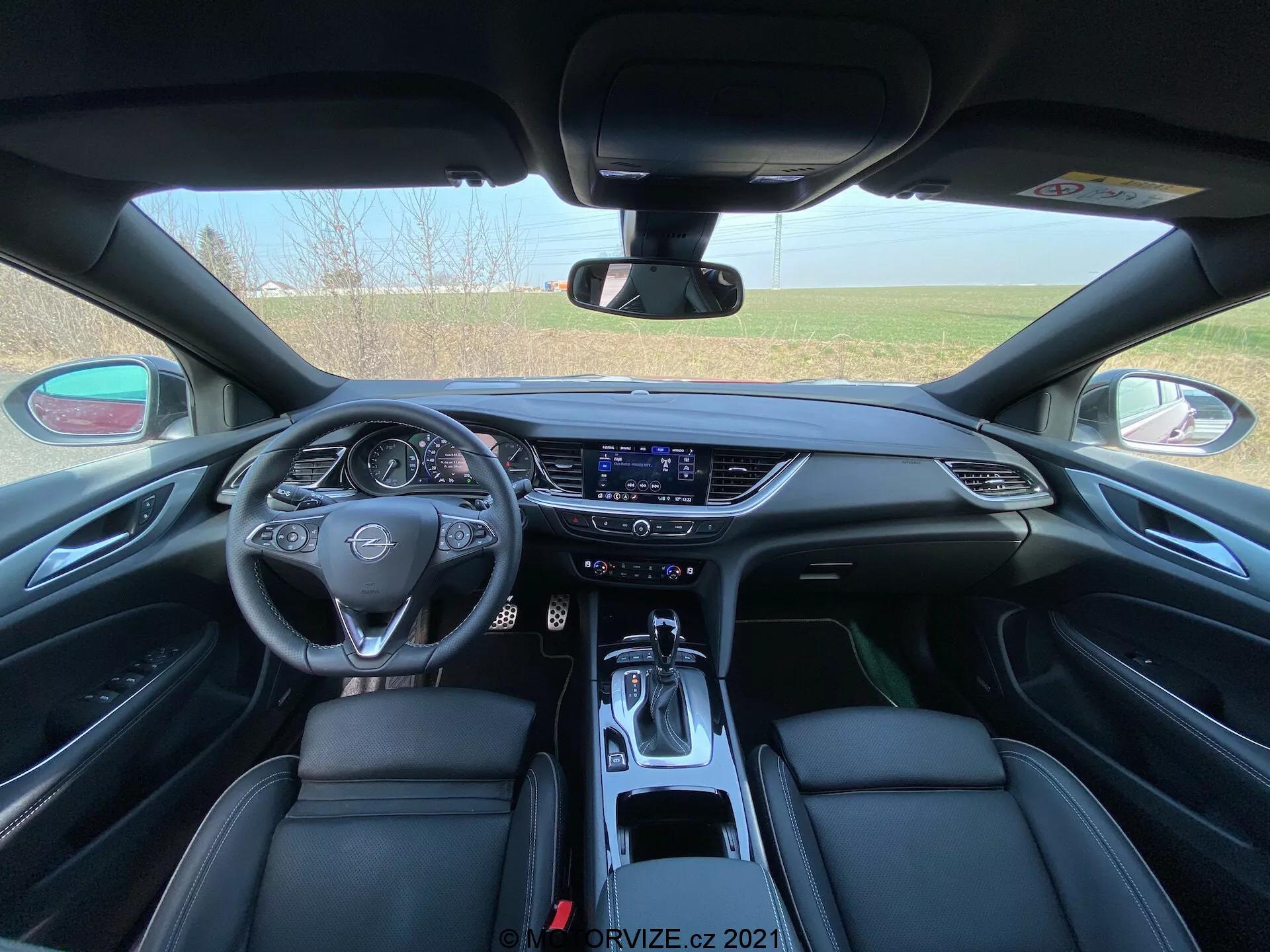 Vue intérieure de l'Opel Insignia GSI Grand Sport 2020 depuis la perspective du siège arrière montrant une vue détaillée du tableau de bord avant. L'image est centrée avec le volant multifonction en cuir avec l'emblème de la marque, un tableau de bord numérique et un écran tactile d'infodivertissement actif. La console centrale avec des bouches d'aération élégantes, des boutons de contrôle du climat et un levier de vitesses au design moderne met en valeur les caractéristiques technologiques. Les sièges avant en cuir noir avec des coutures détaillées ajoutent à l'élégance de l'intérieur de la voiture.
