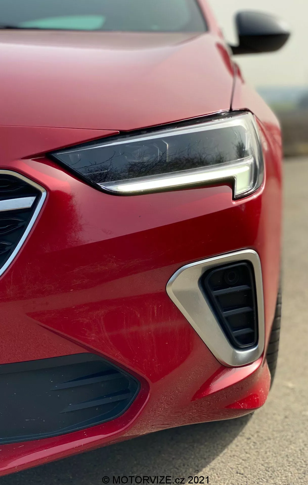 Gros plan sur le coin avant d'une Opel Insignia 2020 rouge moderne (GSI Grand Sport), présentant son phare à LED étroit et élégant, le design de la calandre géométrique, la prise d'air avec accent chromé et le logement possible des phares antibrouillard vue sous un angle bas.