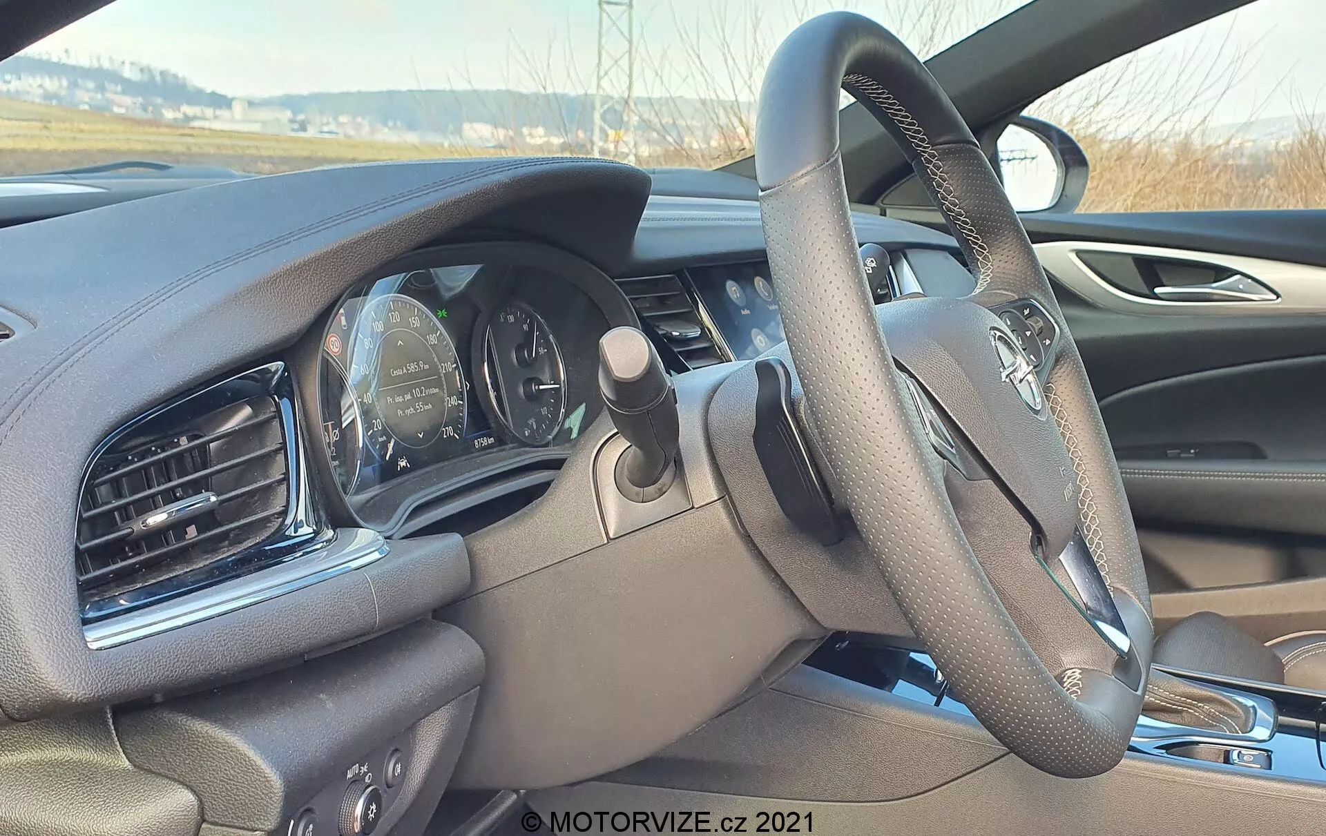 Vue intérieure de l'Opel Insignia 2020 (Insignia-GSI-Grand-Sport) depuis le siège passager, focalisée sur le volant et le tableau de bord. Angle légèrement incliné vers le bas depuis le côté droit, montrant le volant multifonction avec logo flou et le tableau de bord analogique partiellement visible avec le compteur de vitesse et le tachymètre. Tableau de bord avec bouches d'aération et design sculpté, finition en matériau sombre. Aperçu de l'écran d'infodivertissement à droite.
