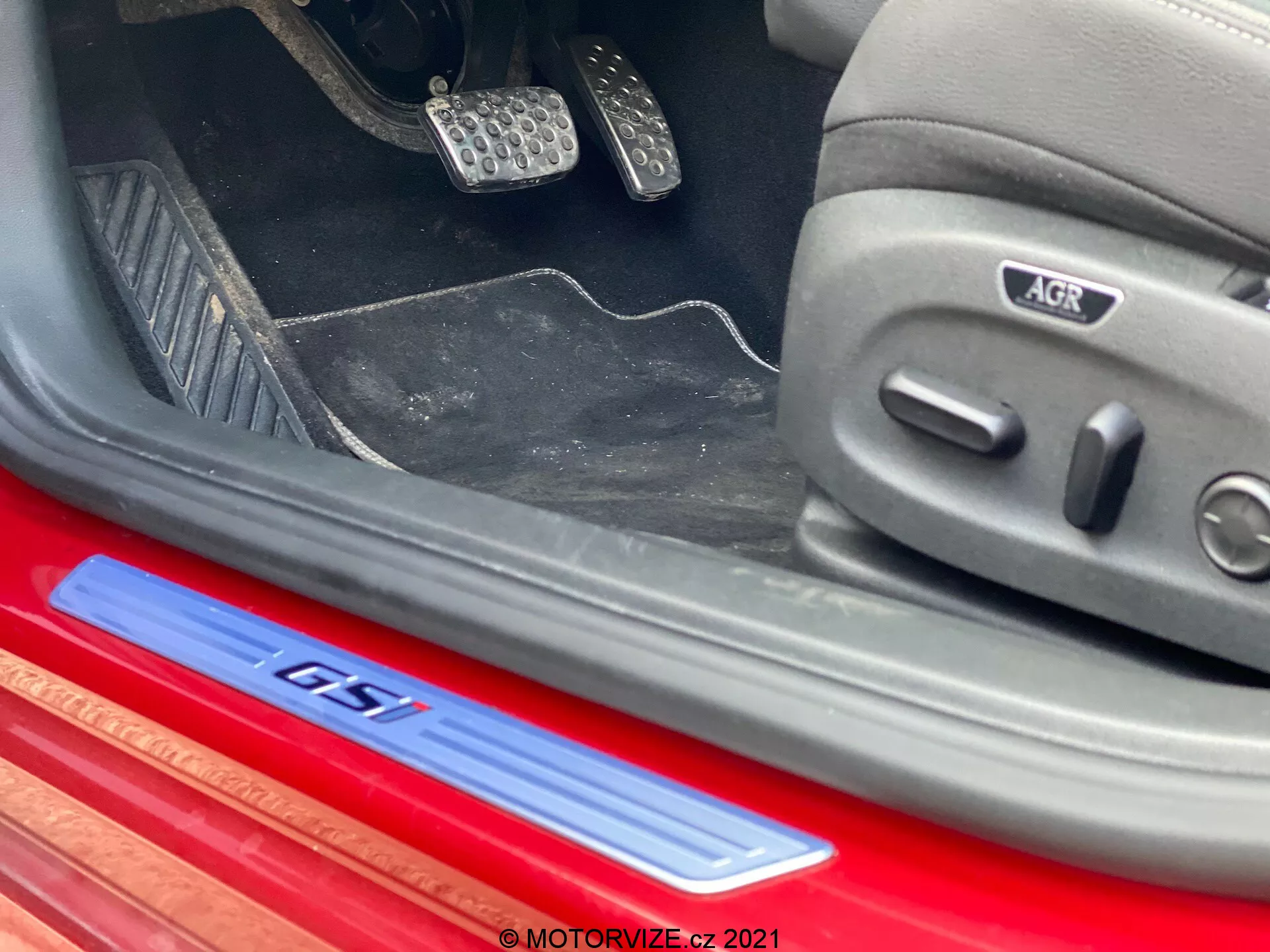 Vue rapprochée en angle de la section inférieure de l'intérieur d'une Opel Insignia GSI Grand Sport 2020. Les caractéristiques visibles incluent une plaque de seuil GTI rouge avec logo argent et bleu sur le seuil de porte en aluminium brossé, des pédales sport en métal avec inserts en caoutchouc, des tapis partiellement visibles et un siège réglable électroniquement avec icônes et badge de certification ergonomique AGR. L'intérieur présente un design sportif et premium avec des tons sombres et des accents rouges et métalliques.