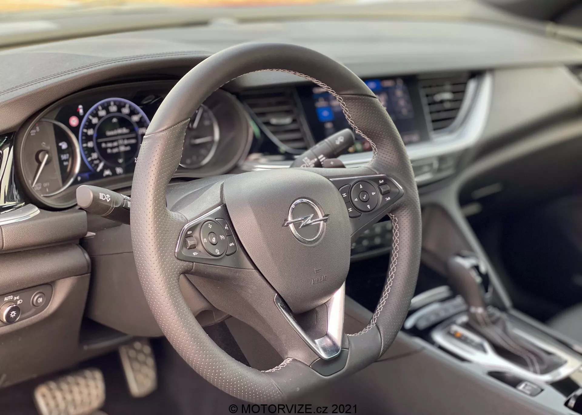 Intérieur de l’Opel Insignia GSi Grand Sport 2020 vu de la perspective du conducteur, mettant en avant le volant centré doté de boutons de commande pour le multimédia et le régulateur de vitesse. Derrière le volant se trouve le tableau de bord avec un compteur de vitesse, un compte-tours et un écran numérique. La console centrale avec les bouches d’aération, un écran d’infodivertissement et les commandes de climatisation est partiellement visible. Le volant en cuir et le design moderne soulignent le confort et la technologie.