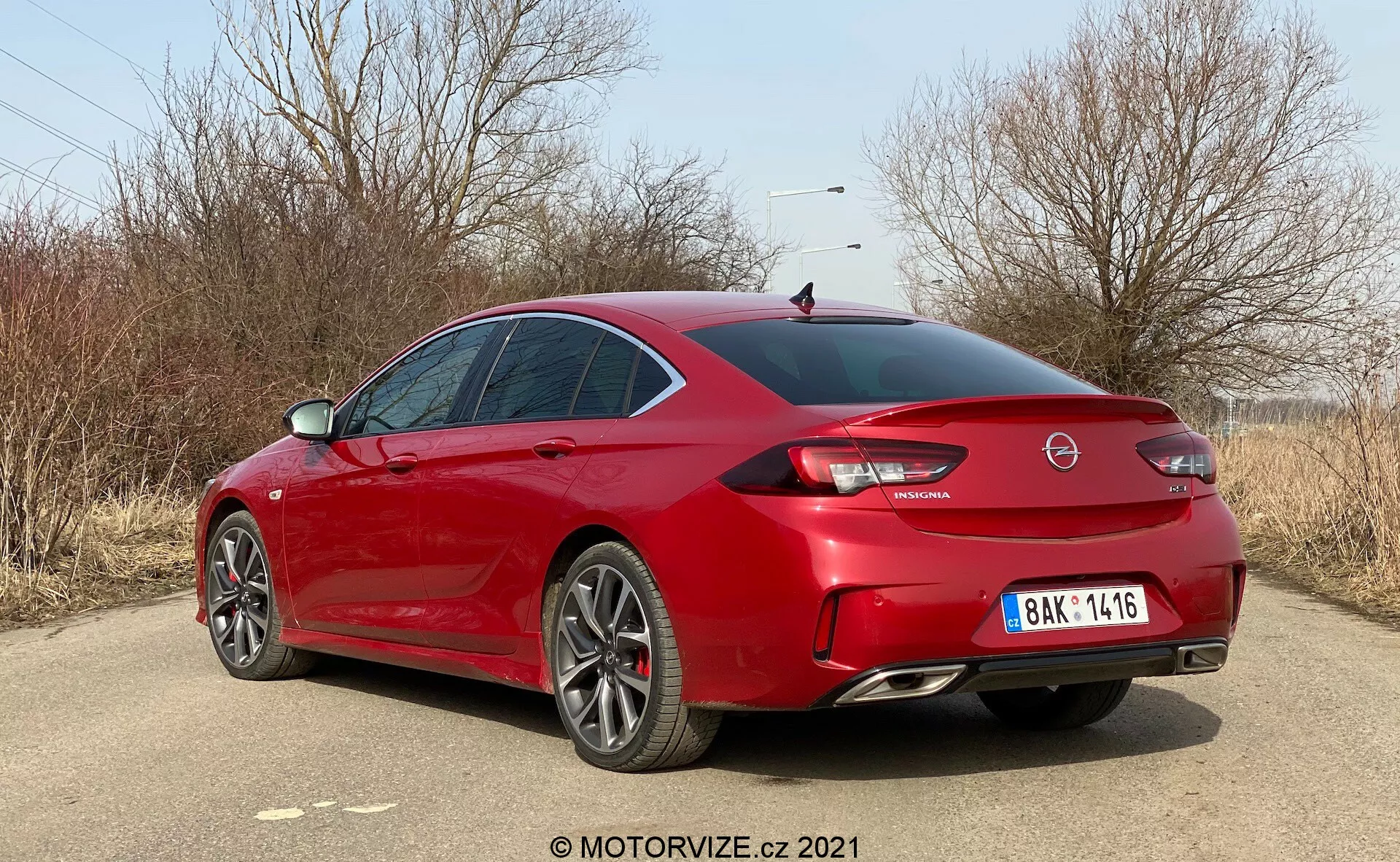 L'image capture une Opel Insignia GSi Grand Sport 2020 rouge sous un angle arrière trois-quarts, en mettant en avant le côté droit et l'arrière du véhicule positionné en diagonale. La photo, prise en plein jour dans un endroit extérieur sec et herbeux avec des arbres nus, représente probablement la fin de l'automne ou l'hiver. La berline moderne, élégante et sportive, présente des lignes de carrosserie acérées, une lunette arrière inclinée, un coffre court, un aileron arrière proéminent, de grandes jantes en alliage à plusieurs branches, des doubles sorties d'échappement et un diffuseur arrière notable. Les feux arrière à LED, le logo central sur le coffre, l'emblème du modèle et la plaque d'immatriculation centrée sont visibles. Les vitres teintées et les rétroviseurs latéraux de la couleur de la carrosserie contribuent à l'allure agressive mais élégante de la voiture.