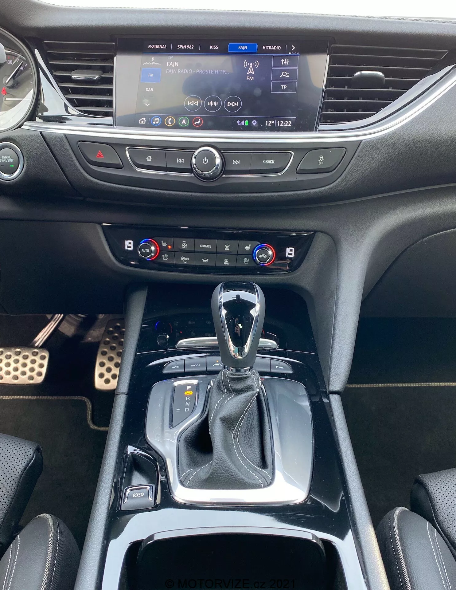 Image de l'intérieur de l'Opel Insignia GSi Grand Sport 2020, montrant l'espace de la cabine avant du point de vue du conducteur ou du passager avant. La photo capture la console centrale du levier de vitesses à l'écran d'infodivertissement. Le levier de vitesses est en position parking, suggérant une transmission automatique, et est entouré d'un garniture noire brillante. À proximité se trouvent des boutons de modes de conduite et un soufflet en cuir autour du levier de vitesses qui ajoute une touche premium. Derrière le levier de vitesses se trouvent deux porte-gobelets et une zone de rangement avec un couvercle rétractable. Au-dessus, les commandes de climatisation avec affichages de température à double zone et boutons physiques. Plus haut, le système d'infodivertissement activé affiche une carte de navigation et d'autres paramètres. Le tableau de bord en plastique au toucher doux intègre sans interruption les bouches d'aération de chaque côté de l'écran d'infodivertissement.