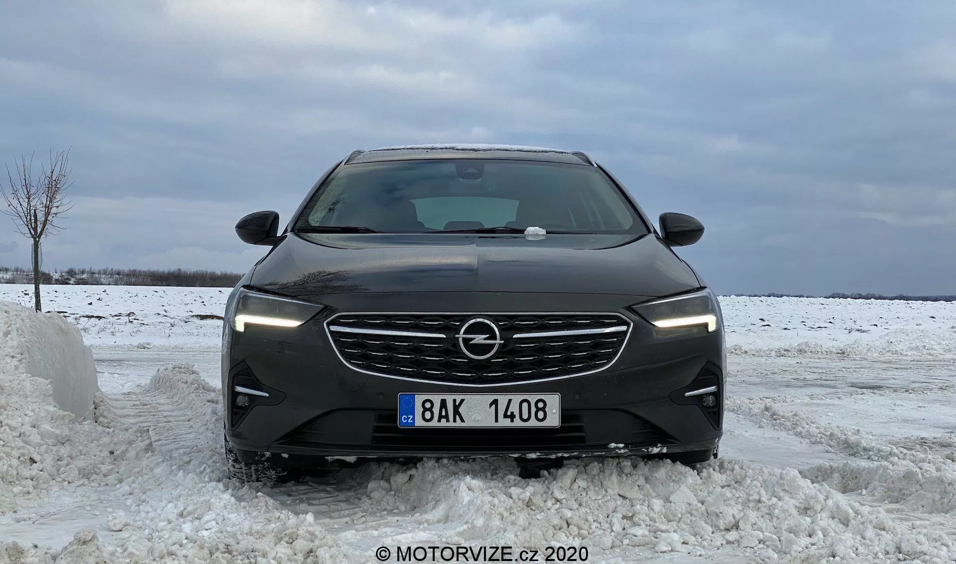 Vue de face d'une Opel Insignia Sports Tourer 2020 sur une route enneigée montrant ses éléments de design modernes tels que les lamelles horizontales sur la grille, les phares LED angulaires, les contours sculptés du capot, le large pare-chocs avant avec des prises d'air intégrées et la plaque d'immatriculation centrale, mettant en avant les conditions hivernales.