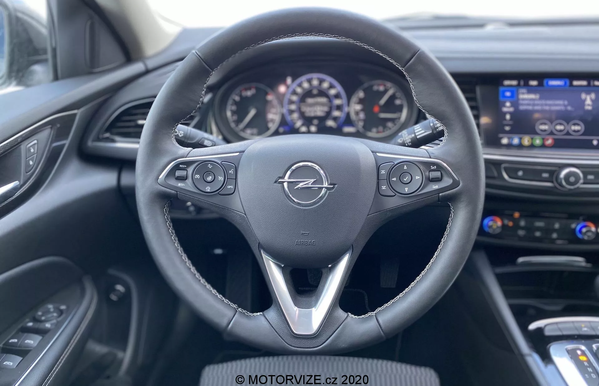 Vue depuis la perspective du siège du conducteur de l'Opel Insignia Sports Tourer 2020, montrant le volant multifonction en cuir avec un design à trois branches, les commandes du système d'infotainment, les réglages du régulateur de vitesse, tableau de bord partiellement visible avec cadrans analogiques et petit écran numérique, écran d'infotainment sur la console centrale, aérations, système de contrôle du climat avec boutons et commandes, tableau de bord de couleur sombre avec motif texturé et panneau de contrôle de la porte du conducteur partiellement visible.