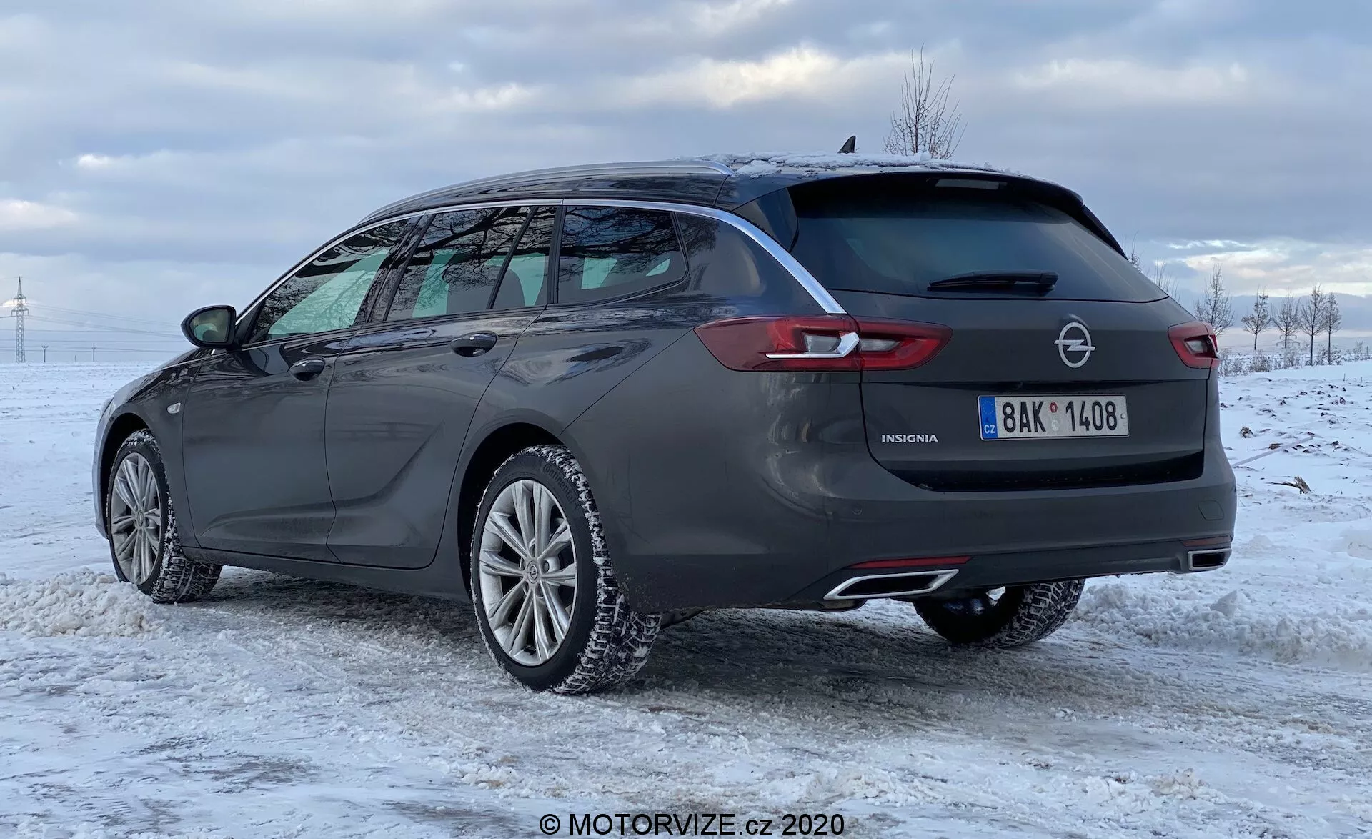 Vue de côté et arrière d'une Opel Insignia Sports Tourer 2020 de couleur sombre dans un paysage enneigé, montrant son design moderne et élégant. La voiture est équipée de jantes en alliage, de rails de toit, de double tuyaux d'échappement, de feux arrière à LED, d'un spoiler arrière, d'un essuie-glace arrière et d'une antenne aileron de requin. Cette image met en avant la grande capacité de chargement, l'esthétique sportive et les fonctionnalités pratiques du modèle contemporain.