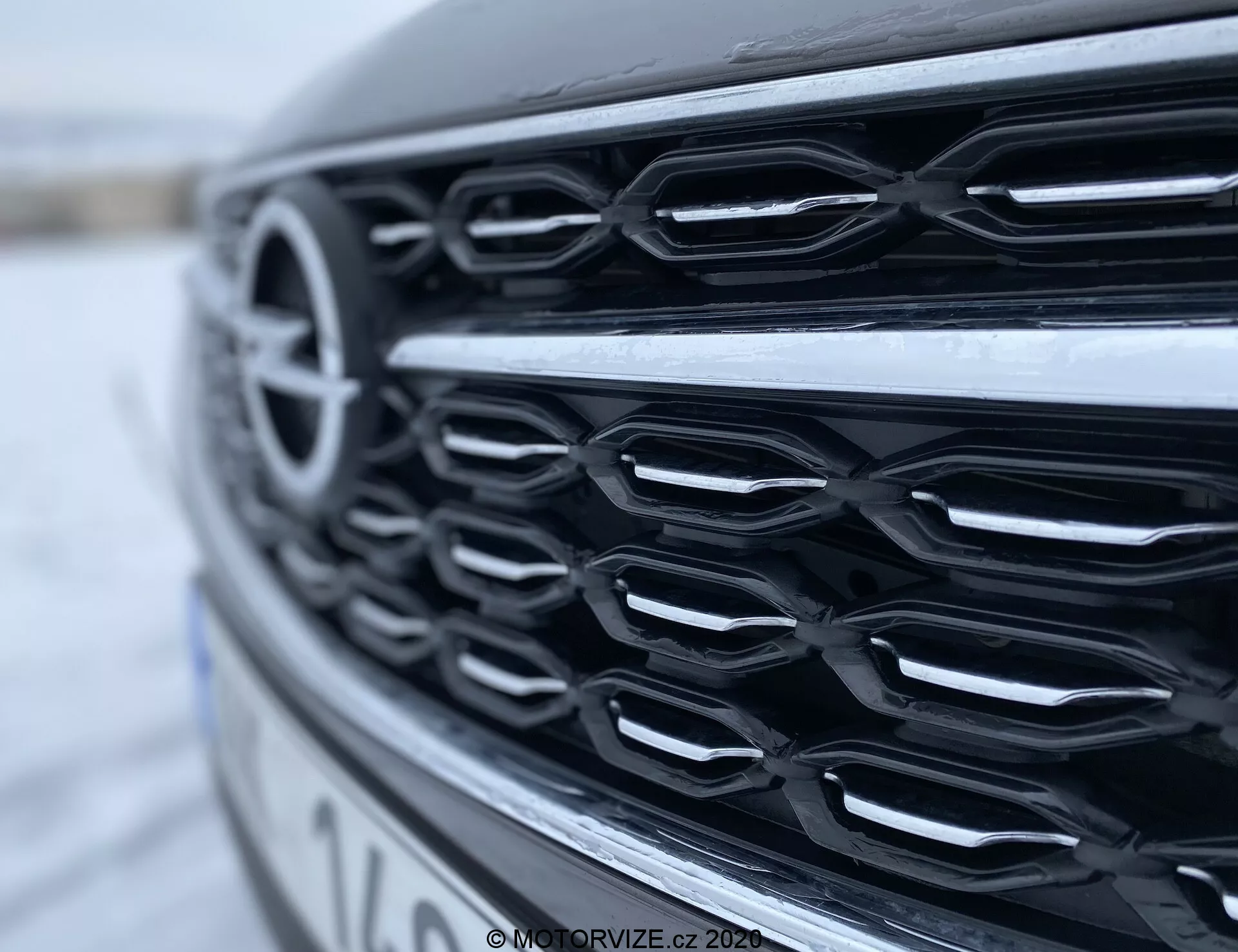 Gros plan en profil latéral de la calandre avant de l'Opel Insignia Sports Tourer 2020, mettant en valeur des lamelles horizontales aspect chromé avec une légère courbure, capturé sous un angle bas. Les reflets sur la finition brillante apparaissant sous un éclairage naturel diffus, mettent en évidence les contours de la calandre et la profondeur, tandis que l'arrière-plan flou accentue son design moderne.