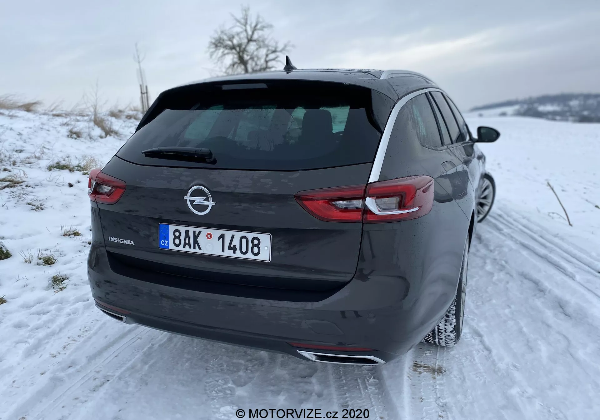 Vue arrière d'une Opel Insignia 2020 moderne hatchback garée sur une route enneigée, montrant le côté arrière droit, le coffre, le pare-chocs et une partie de la porte passager arrière. La voiture est équipée de feux arrière à LED, d'un essuie-glace arrière, d'une antenne aileron de requin sur le toit, ainsi que du nom du modèle et de l'emblème Opel sur le hayon. Plaque d'immatriculation de style européen bien visible.