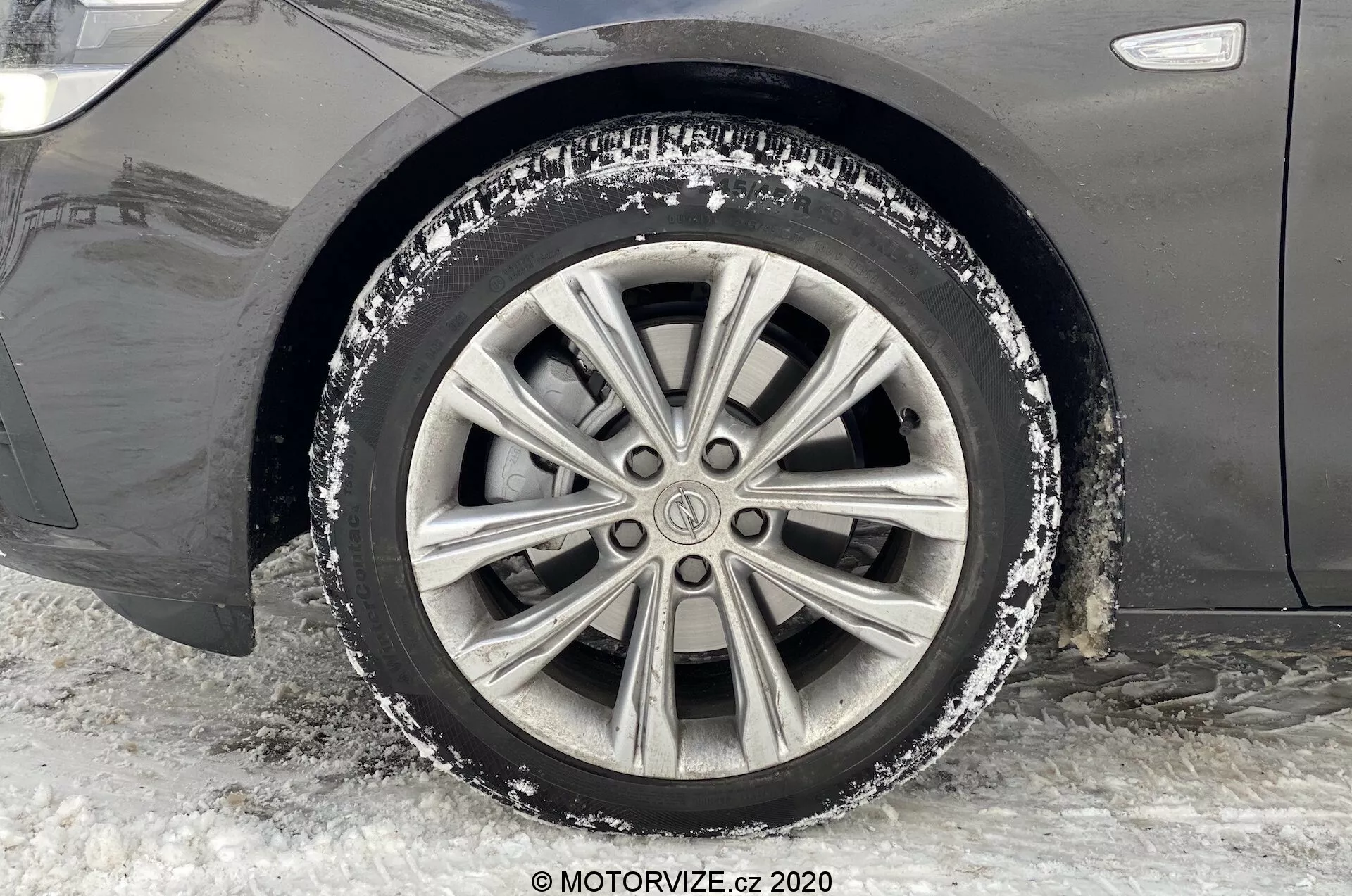 Vue rapprochée de la roue avant et de l'aile du côté passager d'une Opel Insignia 2020 (Insignia Sports Tourer). L'image est prise au niveau du sol, capturant la perspective latérale. La voiture est stationnée sur de la neige et de la glace compactées, indiquant un environnement hivernal. La roue présente une jante en alliage à multiples rayons avec un peu de neige et de sel de voirie, montrant un trajet récent sur des routes hivernales. Des marques d'usure et de la saleté sont visibles sur la jante, reflétant l'utilisation dans des conditions enneigées.