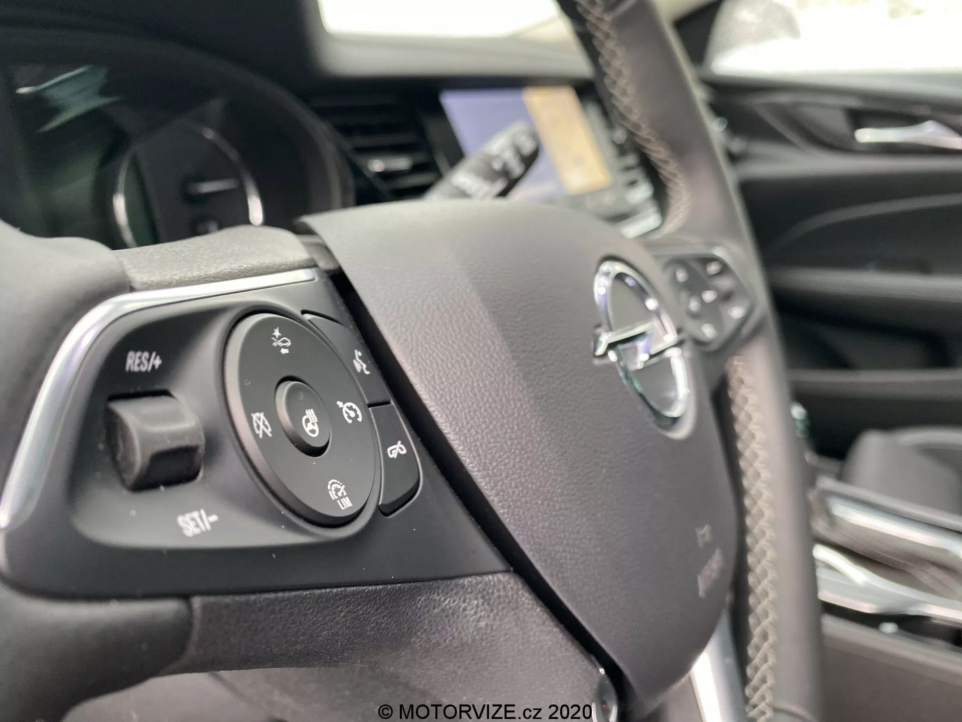 Vue rapprochée du volant d'une Opel Insignia Sports Tourer 2020 du point de vue du conducteur, montrant les commandes intégrées à gauche, y compris le régulateur de vitesse, la commande vocale, les fonctions téléphoniques et les commandes audio. Le volant présente des boutons tactiles pour une utilisation facile et un emblème de fabricant flou au centre, dans un intérieur premium avec une finition en fibre de carbone.