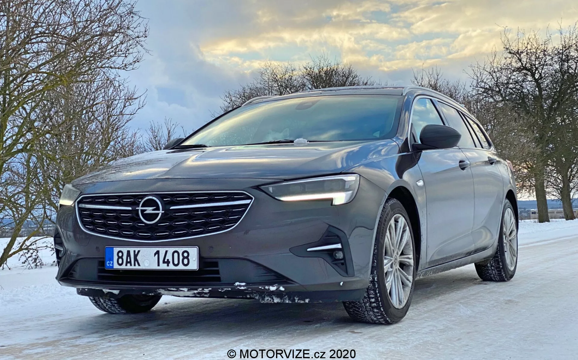 Vue frontale basse d'une Opel Insignia Sports Tourer 2020, avec une calandre avant proéminente au design de grille élégant, un ensemble de phares complexe avec technologie LED avancée, un pare-chocs avant aérodynamique, une roue en alliage à multiples rayons et un clignotant intégré dans le rétroviseur latéral. Le véhicule est sur une route enneigée, avec des arbres et un ciel nuageux en arrière-plan, mettant en avant le design moderne de la voiture et les conditions de conduite hivernales.