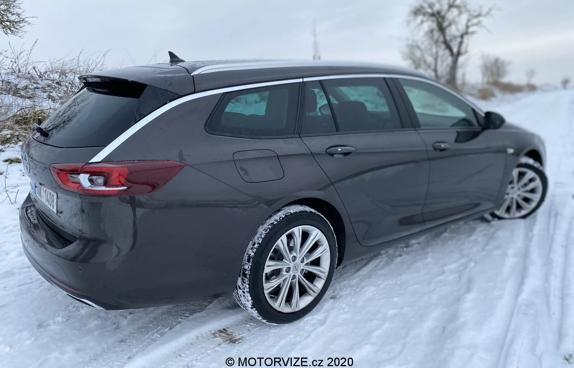 L'Opel Insignia Sports Tourer 2020, un élégant break moderne, est montré dans un environnement extérieur hivernal sous un angle arrière trois quarts, mettant en valeur son profil aérodynamique, son hayon arrière, ses feux arrière, son pare-chocs arrière, ses rétroviseurs latéraux, ses roues en alliage et ses lignes distinctives de carrosserie. Le coffre ouvert suggère un grand espace de chargement et l'arrière-plan enneigé indique la capacité du véhicule à affronter des conditions hivernales. Les feux arrière arborent un design LED horizontal enveloppant les côtés, la plaque d'immatriculation est floue et l'image comporte un filigrane MOTORVIZE.cz 2020.