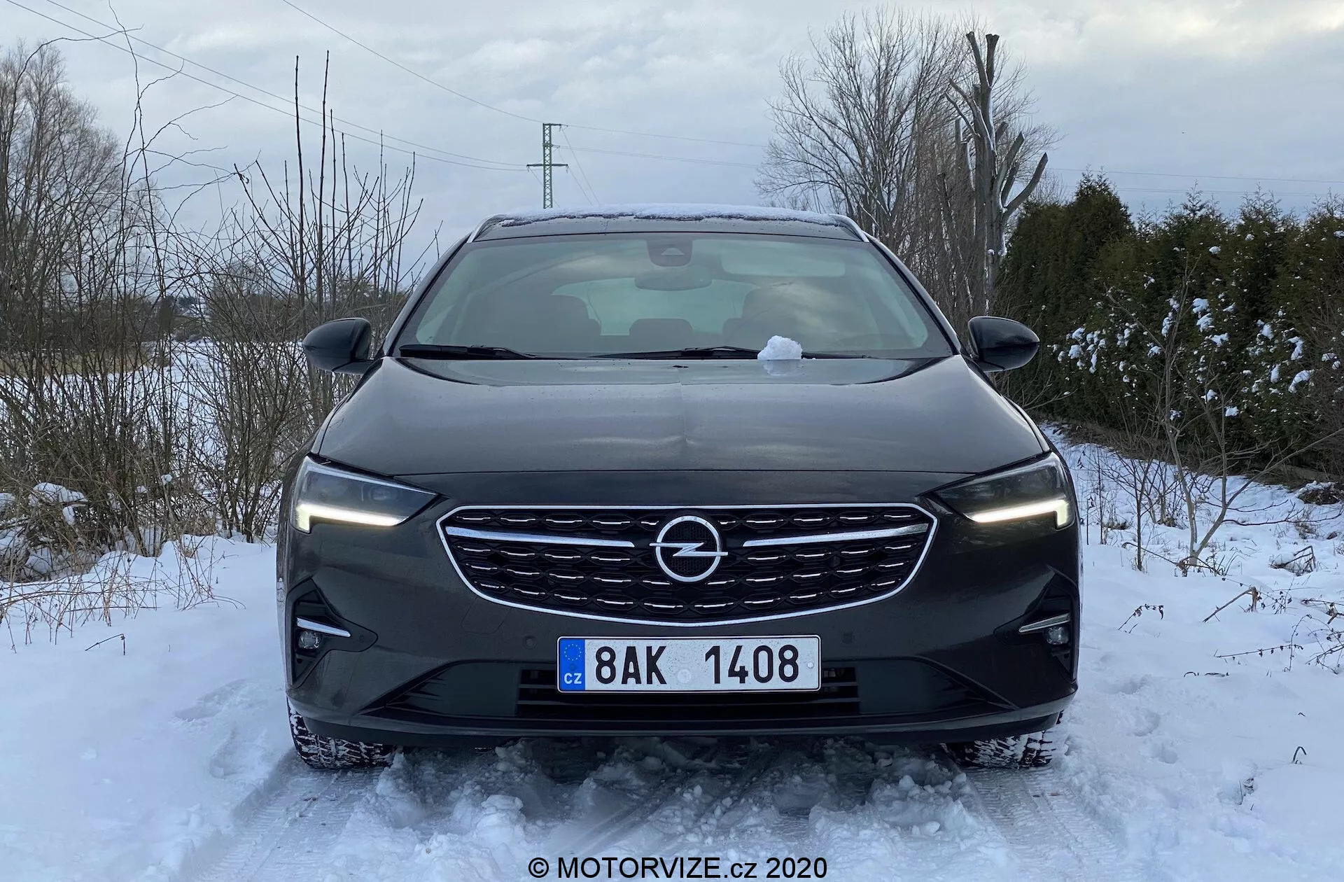 Vue frontale d'une Opel Insignia Sports Tourer 2020 stationnée dans un environnement enneigé. On aperçoit le capot, la calandre, les phares et le pare-chocs avant, avec de la neige au sol et sur la voiture. La calandre présente un badge central proéminent et un design à lamelles horizontales. Les phares élégants s'étirent horizontalement, tandis que le pare-chocs inclut des feux antibrouillard, des prises d'air et des capteurs de stationnement. Les rétroviseurs sont déployés, indiquant que la voiture est prête à être conduite. La lumière naturelle nuageuse offre un éclairage uniforme sans ombres marquées.