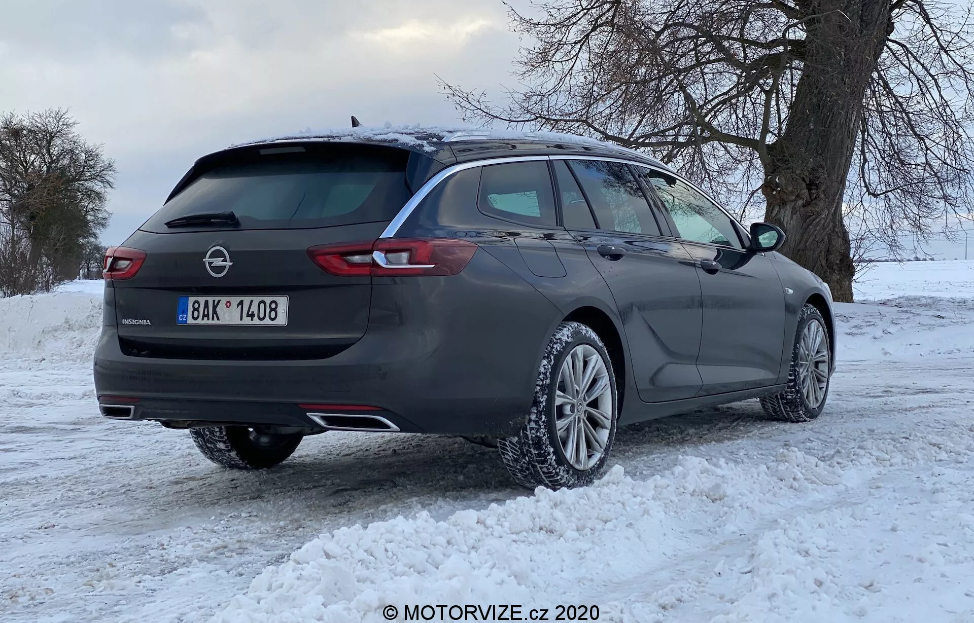 Break Opel Insignia 2020 gris foncé avec des jantes en alliage multi-bâtons sur sol enneigé, vue arrière trois quarts montrant des feux arrière à LED horizontaux, barres de toit, vitrages teintés et badges indiquant la marque et le modèle, avec un arrière-plan hivernal d'arbres sans feuilles et un ciel nuageux.