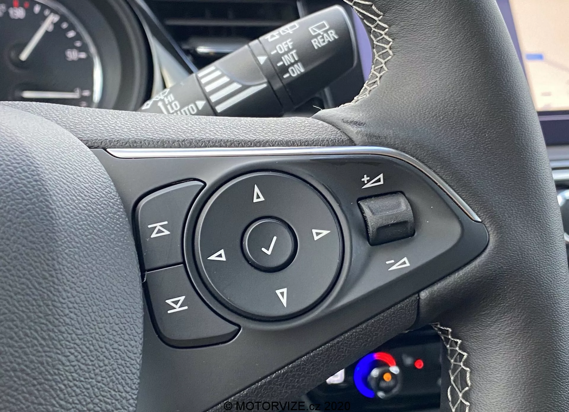 Vue rapprochée des commandes du volant côté gauche d'un véhicule moderne, avec revêtement en cuir avec coutures visibles, un pavé multidirectionnel avec un bouton de confirmation central, des boutons pour les commandes vocales, la gestion des appels téléphoniques, le contrôle du volume et un possible bouton pour la sélection des médias/pistes. Commandes visibles pour les feux de croisement et feu arrière de brouillard, palettes de changement de vitesse et indice d'un affichage digital du tableau de bord en arrière-plan flou, mettant en évidence la technologie moderne des véhicules, les caractéristiques intérieures et les attributs de luxe.