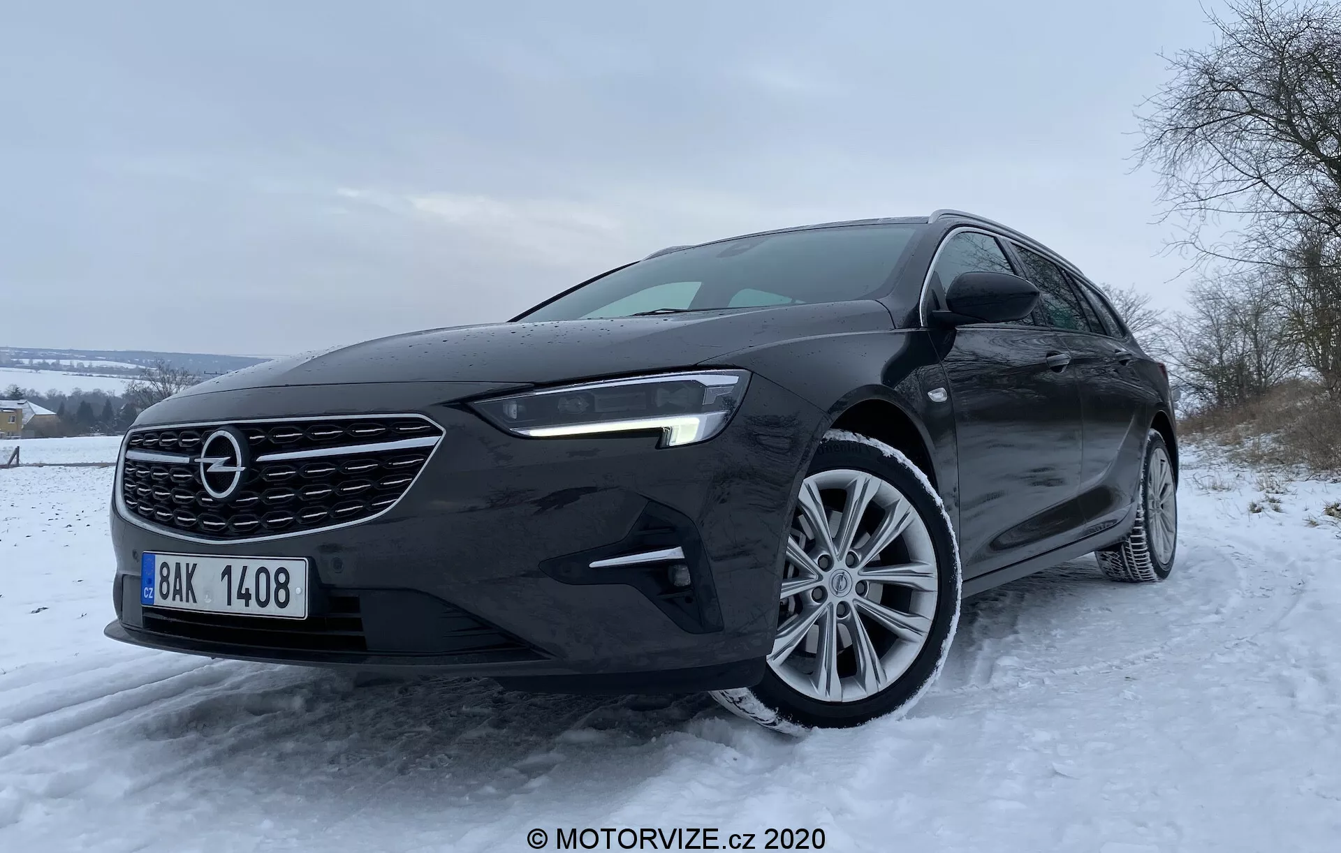 L'image montre une berline Opel Insignia Sports Tourer 2020 vue de bas et de face, mettant en valeur sa calandre distinctive, son pare-chocs avant, ses feux de jour à LED et ses jantes en alliage, sur un fond hivernal avec des arbres enneigés et un ciel couvert.