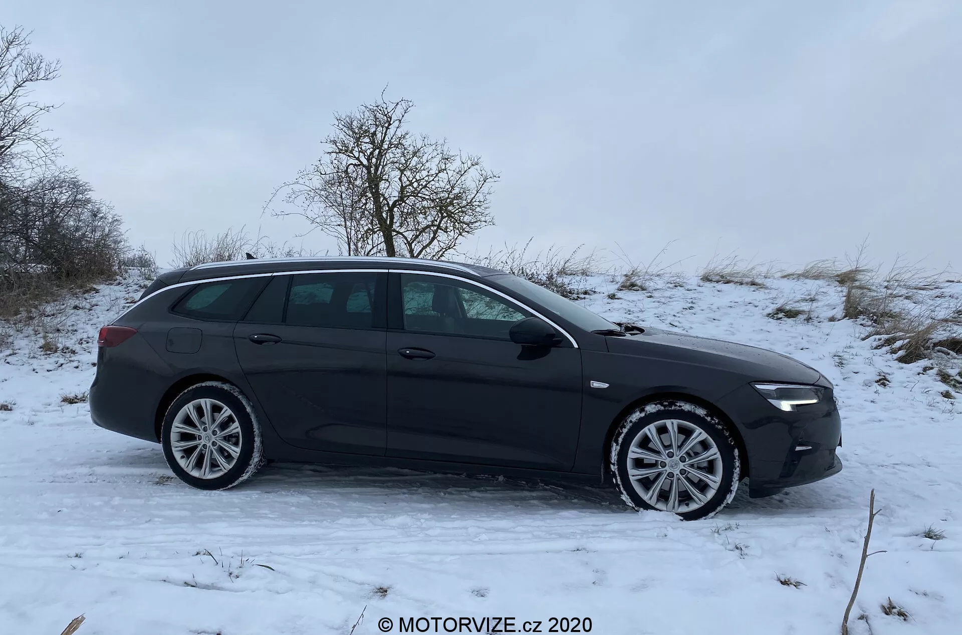 Image d'un break noir Opel Insignia 2020 garé sur une légère inclinaison avec un sol couvert de neige, vue de profil latéral. Le côté gauche entier du véhicule est visible, y compris les jantes en alliage à rayons multiples, les poignées des quatre portes, les rétroviseurs extérieurs, les vitres et les barres de toit. Le design moderne de la voiture présente des lignes de contour lisses et une ligne de toit élégante, suggérant une utilité pour le transport d'objets tels que des porte-bagages ou des équipements sportifs.
