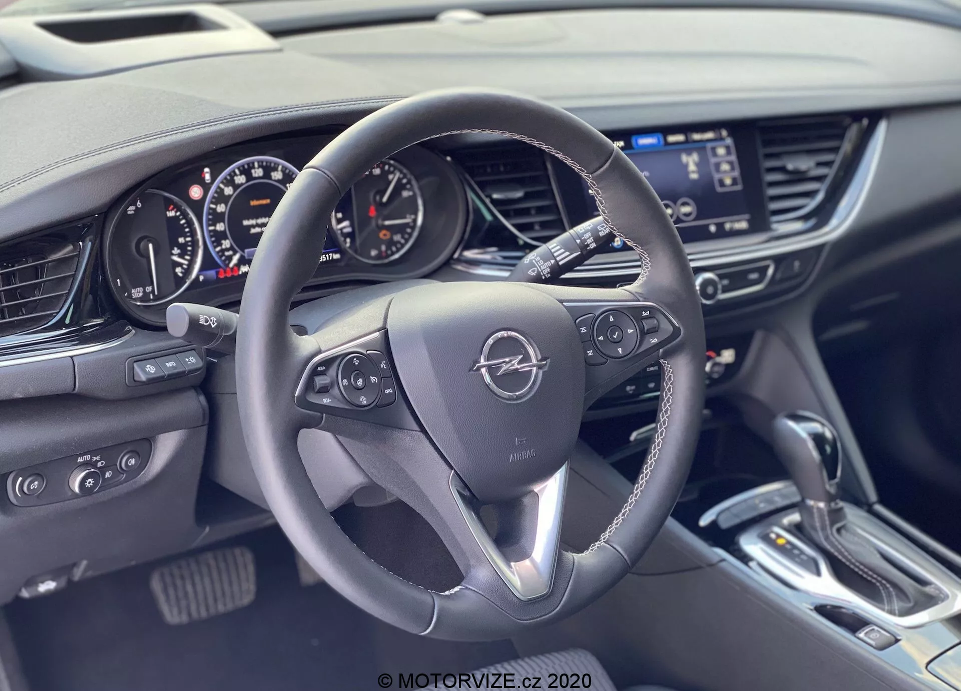 Vue intérieure d'une Opel Insignia Sports Tourer 2020 depuis les sièges arrière, légèrement décalée du côté du conducteur, se concentrant sur le volant et le tableau de bord. Le volant à trois branches présente des accents métalliques et des boutons de commande pour l'infodivertissement et les informations du véhicule. L'emblème est centré bien en vue. Le tableau de bord derrière est une combinaison analogique-numérique avec un compteur de vitesse, un compte-tours et des écrans numériques. À droite, la console centrale abrite un écran d'infodivertissement, des commandes de climatisation et un levier de vitesses moderne. L'intérieur comprend du cuir, des accents métalliques et des matériaux doux au toucher, montrant un design moderne orienté vers la technologie.