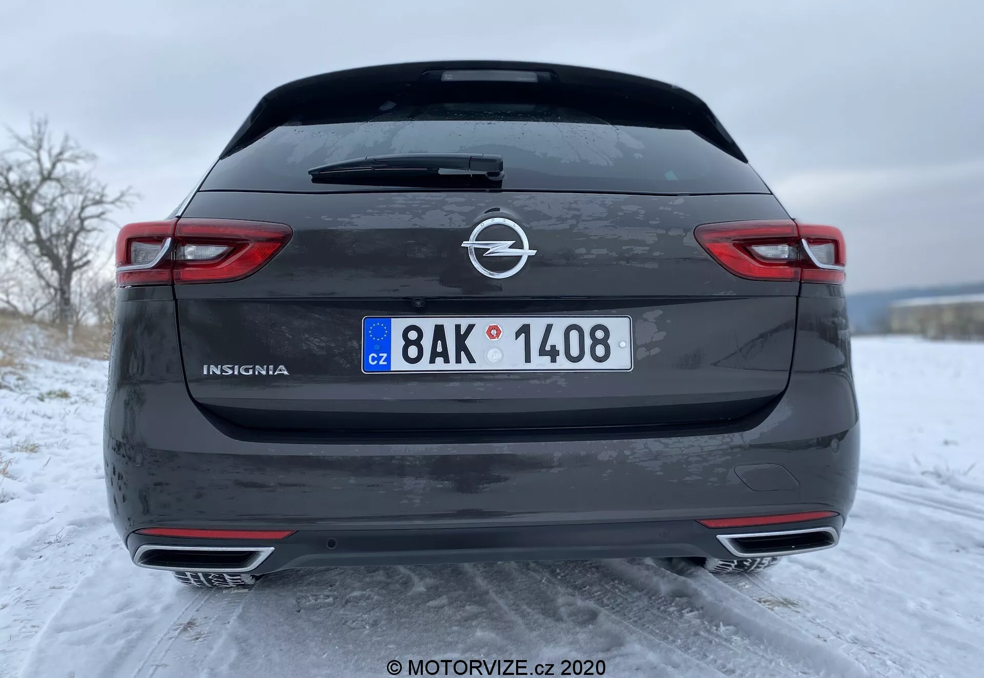 Vue arrière de l'Opel Insignia Sports Tourer 2020 en gris foncé ou noir, présentant le design détaillé avec des feux arrière horizontaux allongés, des doubles sorties d'échappement et des capteurs de stationnement visibles sur le pare-chocs inférieur. La photo met en avant les caractéristiques sportives du véhicule, la plaque d'immatriculation européenne et l'environnement hivernal enneigé.