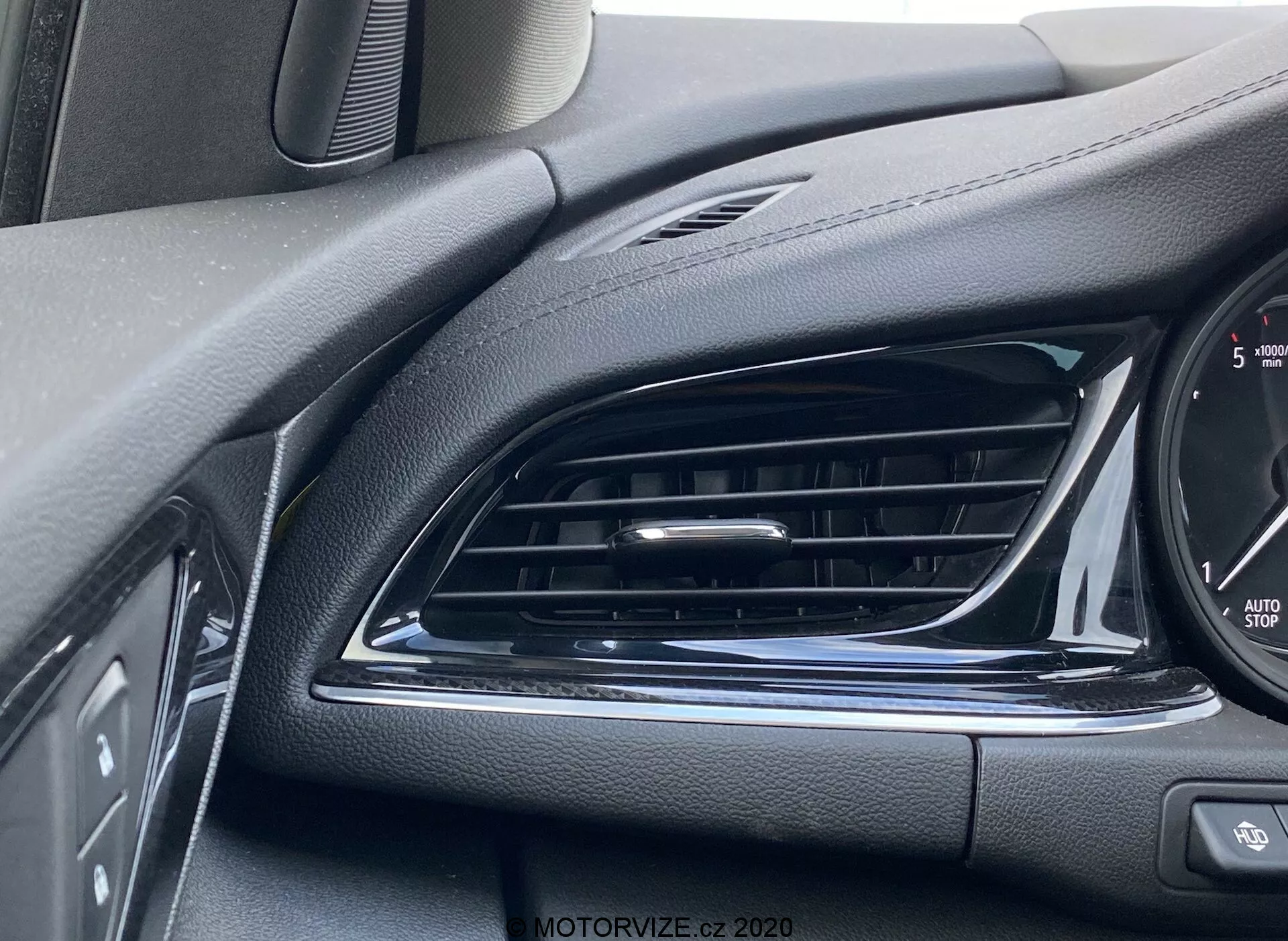 Vue rapprochée de l'intérieur du tableau de bord de l'Opel Insignia Sports Tourer 2020. Comprend une bouche d'aération noire avec des lames horizontales, des surfaces texturées type cuir pour l'esthétique et anti-reflet, un accent chromé brillant, panneau de commandes partiellement visible, bord du tableau de bord avec incréments de vitesse et texte 'Auto Stop' indiquant un système de démarrage et arrêt automatique. Prise de vue angulaire depuis le côté conducteur focalisée sur la bouche d'aération et les détails environnants.