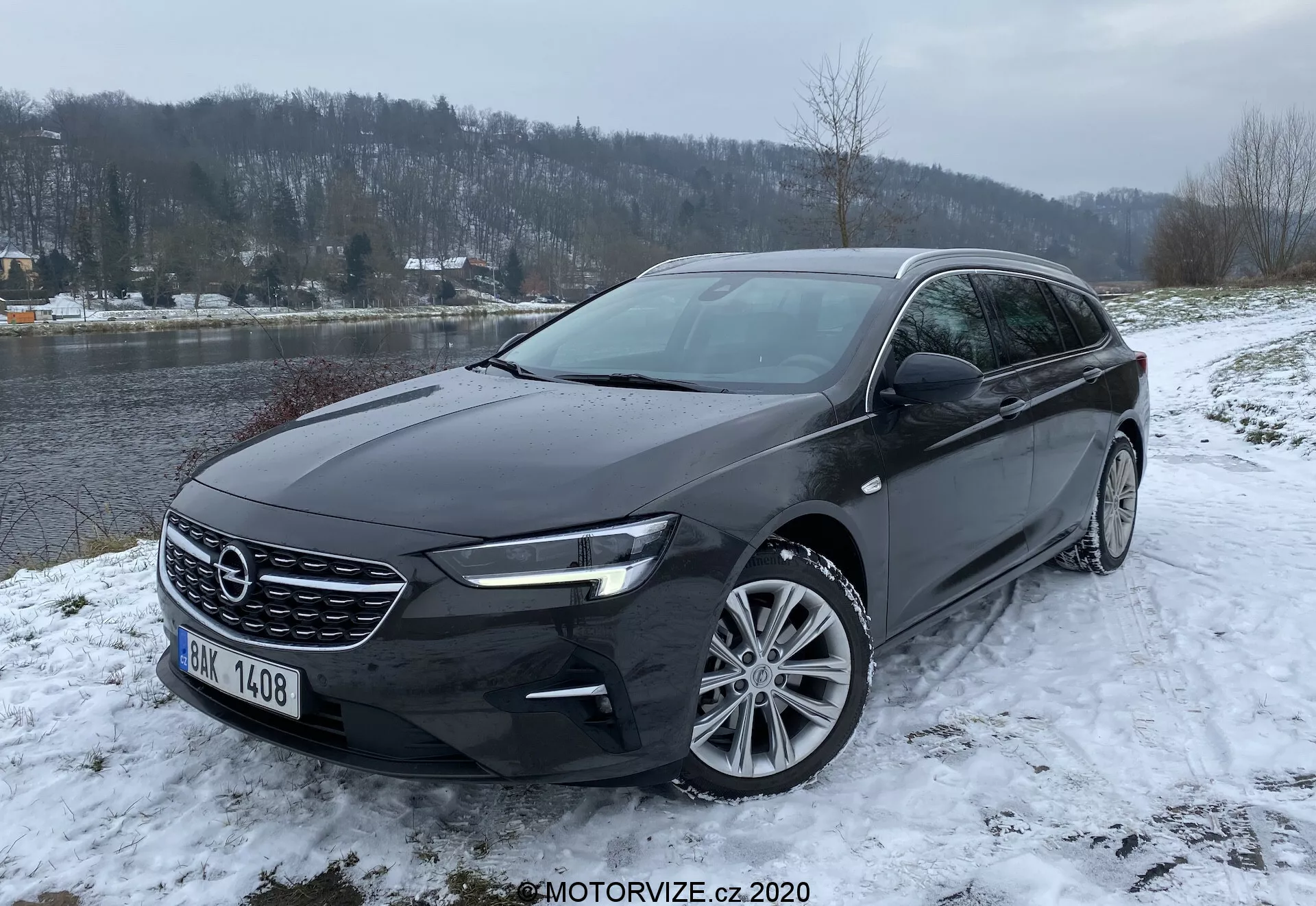 Opel Insignia Sports Tourer 2020 dans un décor hivernal, photographié en vue frontale trois-quarts montrant sa calandre avant proéminente, ses phares LED élégants, ses jantes en alliage multi-rayons et sa couleur extérieure foncée, dans un paysage enneigé avec une rivière et des arbres en arrière-plan.