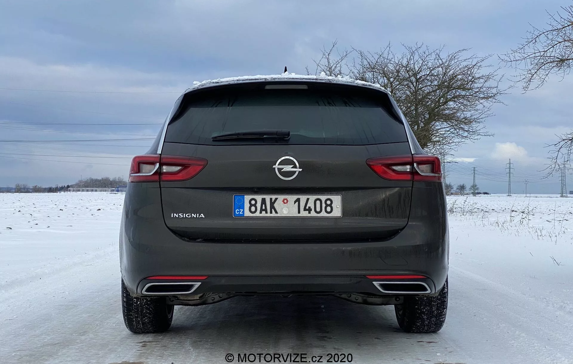 Vue arrière d'une Opel Insignia Sports Tourer 2020 stationnée sur une route couverte de neige. Dotée d'une finition gris métallique, du logo Opel, du nom de modèle 'Insignia', d'une plaque d'immatriculation de style européen, d'un dégivreur arrière avec essuie-glace, d'embouts d'échappement doubles et d'un arrière-plan hivernal avec un champ enneigé et des arbres dénudés sous un ciel nuageux.