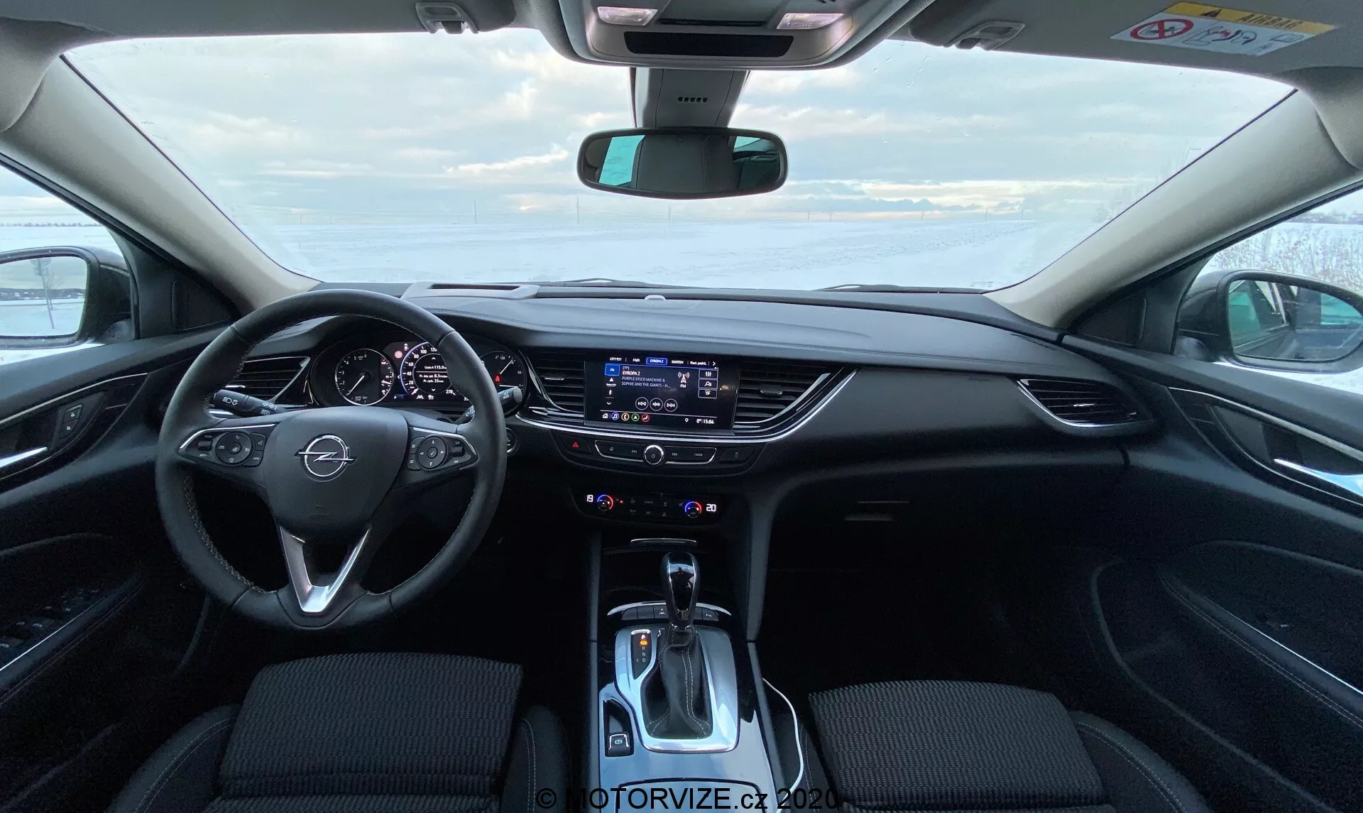 Vue intérieure d'une Opel Insignia Sports Tourer 2020 moderne depuis la perspective du siège du conducteur, présentant le volant avec boutons de commande multimédia intégrés, un tableau de bord partiellement visible et élégant, un système d'infodivertissement à écran tactile large et intégré au tableau de bord, des commandes de climatisation bi-zone, un levier de vitesses moderne et une sellerie en tissu sombre au premier plan.
