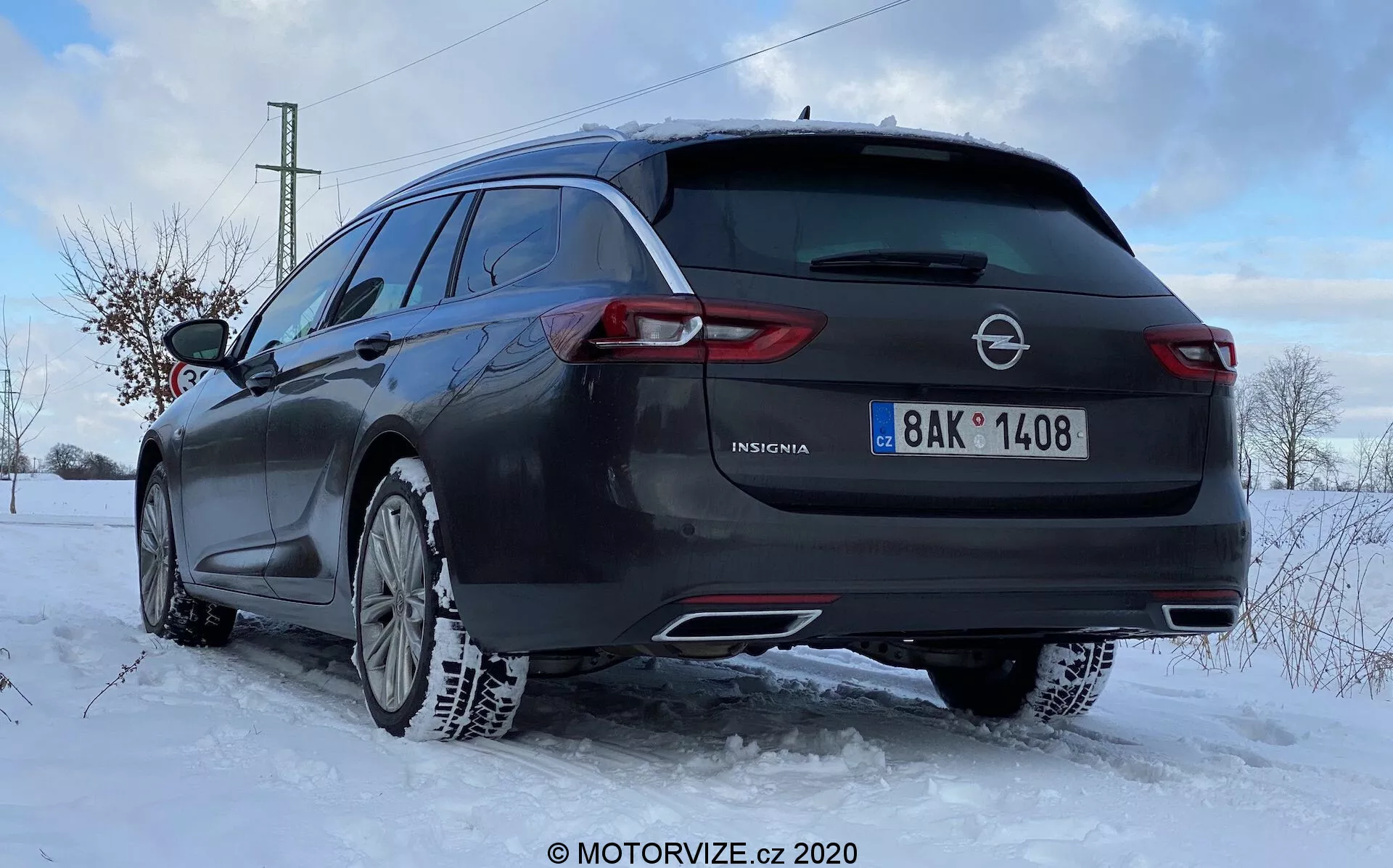 Angle arrière d'une Opel Insignia Sports Tourer 2020 pris à une hauteur basse proche des roues du véhicule. L'image montre le coin arrière droit, affichant le côté droit et l'arrière complet de la voiture. Le hayon est visible avec le logo Opel, le nom du modèle 'Insignia' et une plaque d'immatriculation centrée. Un essuie-glace arrière est en haut de la lunette arrière et les feux arrière enveloppants s'étendent sur les côtés. Le système d'échappement double suggère des performances, et la voiture est équipée de pneus neige, indiquant des conditions hivernales. Il y a de la neige sur le sol et la moitié inférieure du véhicule. La lumière suggère l'aube ou le crépuscule avec un paysage enneigé et légèrement couvert. Les éléments de design modernes incluent des feux arrière à LED, des capteurs de stationnement potentiels, une antenne radio montée sur le toit et une possible caméra arrière.