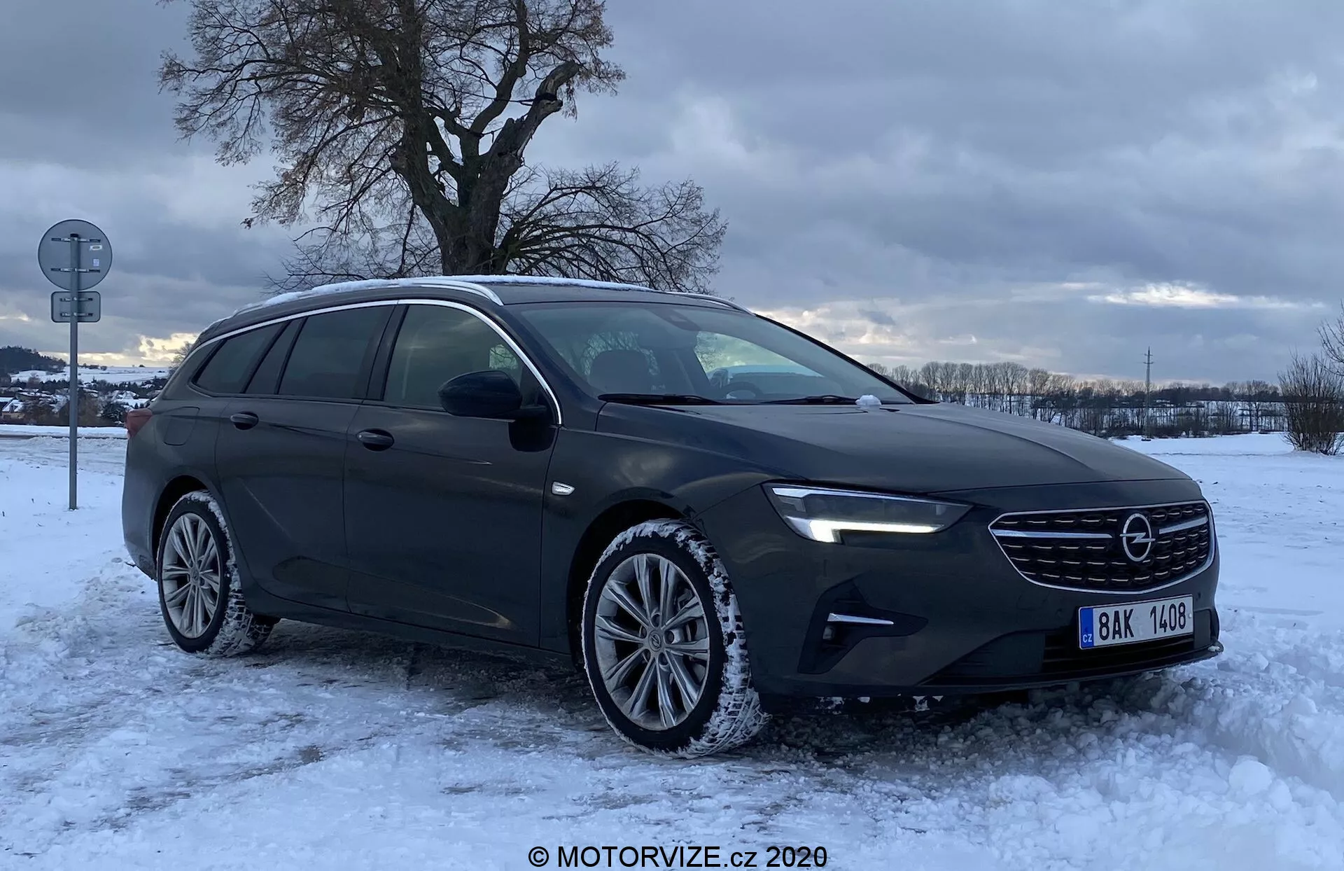 L'image montre une Opel Insignia Sports Tourer 2020 photographiée de profil avant trois quarts. L'angle met clairement en évidence la calandre avant avec l'emblème de la marque, les phares élégants pouvant inclure une technologie LED, les contours du capot et le côté conducteur, y compris les portes avant et arrière et les rétroviseurs latéraux. La forme allongée et la ligne de toit étendue du break mettent en avant son espace de chargement plus grand. Dans un paysage hivernal avec de la neige au sol et un ciel couvert, la voiture est garée sur un sol enneigé avec des traces de pneus visibles. Le pare-chocs avant comprend des feux antibrouillard et les roues en alliage sont équipées de pneus d'hiver. La finition métallisée sombre de la voiture et les vitres réfléchissantes suggèrent un traitement pour la confidentialité ou la réduction des UV. Un clignotant latéral intégré dans le boîtier du rétroviseur améliore la sécurité en signalant les virages ou les changements de voie.