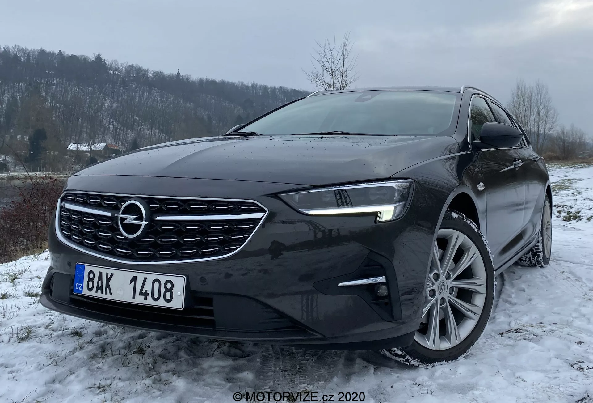 L'image présente un Opel Insignia Sports Tourer 2020 noir capturé sous un angle de trois quarts avant, mettant en valeur le côté gauche du véhicule. Cette perspective inclut le pare-chocs avant, la calandre avec un logo proéminent, le capot, le phare gauche avec une technologie LED avancée, une partie du pare-brise ainsi que la roue et la porte avant du côté conducteur. Les jantes en alliage à plusieurs branches et le rétroviseur latéral avec clignotant intégré sont visibles. La photo est prise d'un angle légèrement plongeant dans un paysage hivernal avec une légère chute de neige ou du givre sur le sol et un ciel couvert, la lumière ambiante suffisante met en avant le design et les éventuelles caractéristiques aérodynamiques et de refroidissement des prises d'air du pare-chocs avant.