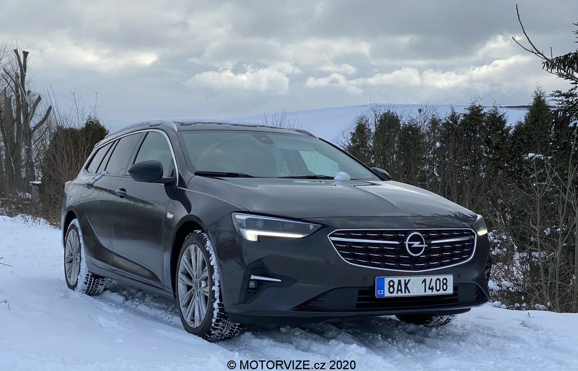 Opel Insignia 2020 Sports Tourer, un break moderne, garé sur un terrain enneigé avec un décor d'arbres hivernaux et un ciel partiellement nuageux. La vue de trois quarts avant montre la calandre élégante du véhicule avec le logo du fabricant, les phares LED et les lignes aérodynamiques sur le capot. Les jantes en alliage à multi-branches montées avec des pneus d'hiver adaptés aux conditions neigeuses sont visibles. La couleur foncée de la voiture, probablement graphite ou gris foncé avec une finition brillante, reflète la lumière naturelle, soulignant un design axé sur la praticité et le confort plutôt que sur les attributs sportifs.