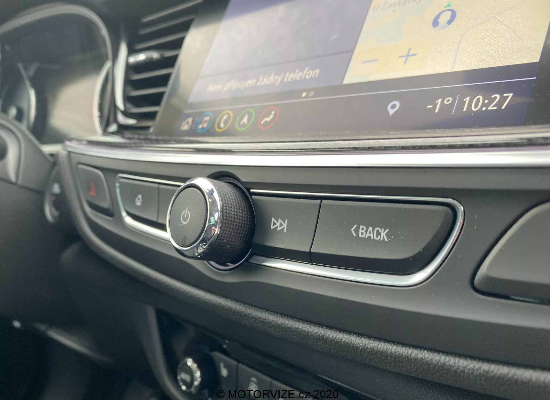 Vue rapprochée de l'intérieur de la Opel Insignia Sports Tourer 2020 depuis le côté conducteur, mettant en avant le bouton de commande rotatif, les boutons noirs mats pour le contrôle audio, et un écran d'infodivertissement flou affichant une interface graphique avec l'heure et la température.