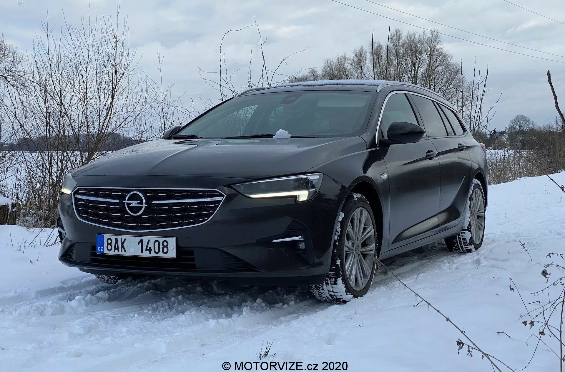 Angle bas trois-quarts avant d'une Opel Insignia Sports Tourer 2020, présentant la calandre avant avec des lamelles horizontales et le logo de la marque en position centrale, des phares modernes et élégants avec feux de jour à LED, un capot profilé, un pare-chocs avant avec prises d'air et probablement des capteurs de stationnement, des roues en alliage et des pneus d'hiver. La voiture, stationnée sur un terrain enneigé, photographiée en plein jour, met en lumière ses capacités de conduite hivernale et sa posture dynamique.