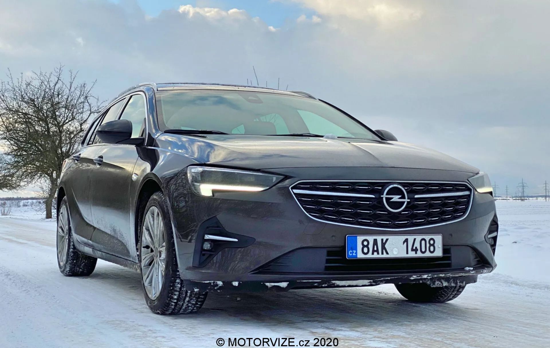 Image d'une voiture moderne (Opel Insignia 2020 Sports Tourer) vue sous un angle de trois quarts avant bas, mettant en avant sa grille frontale proéminente avec l'emblème de la marque, motif horizontal distinctif, phares LED avec feux de jour, capot élégant avec des contours aérodynamiques, jantes en alliage avec pneus d'hiver, rétroviseurs latéraux aérodynamiques avec clignotants intégrés, garnitures de fenêtres chromées, ligne de caractère visible à travers les poignées de porte, pare-brise incliné, couleur sombre brillante, environnement de route enneigée, plaque d'immatriculation avant visible, suggérant des fonctionnalités technologiques avancées.